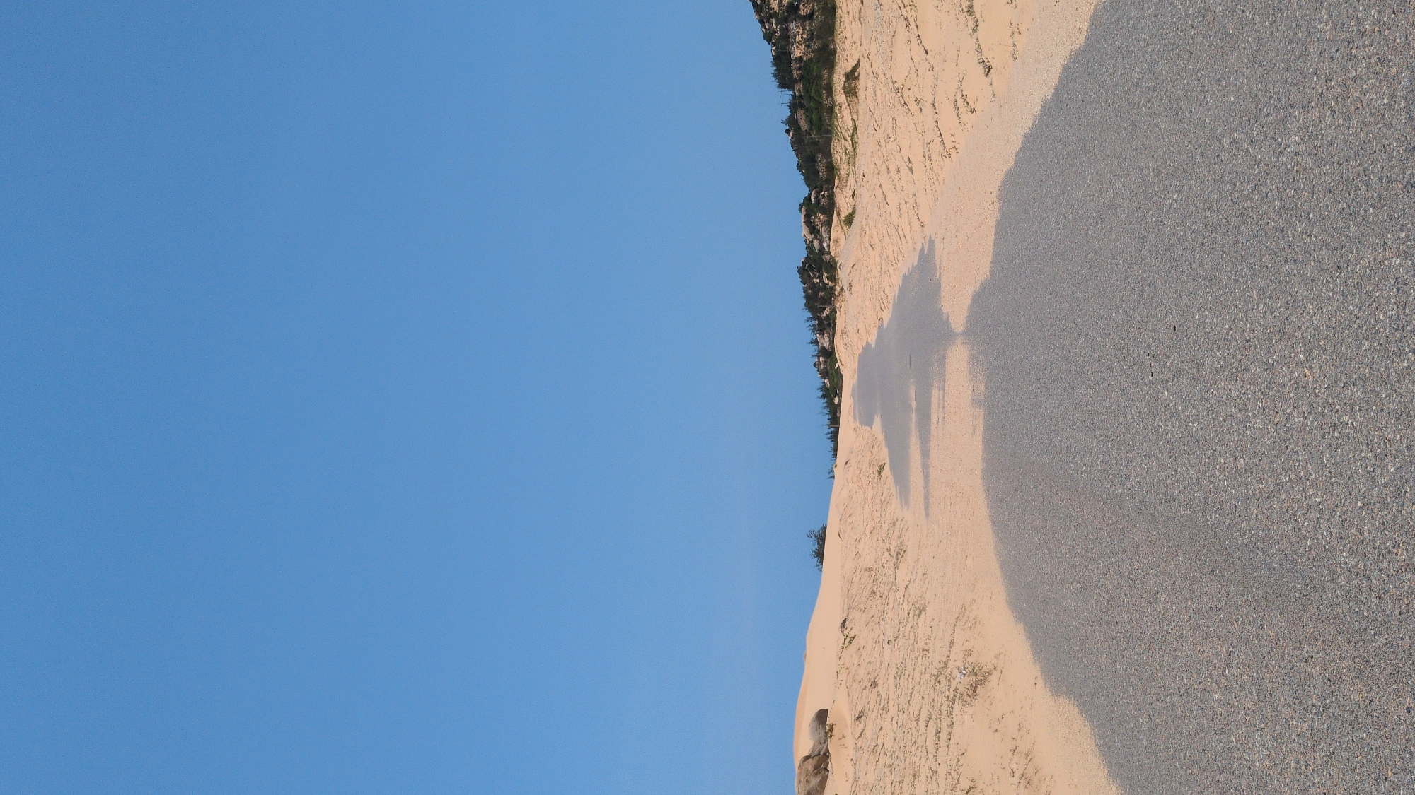 The image depicts a scene in Phuoc Dinh, Vietnam. A landscape consisting of a blue sky and an adjacent area of sand and gravel is visible. Some sparse vegetation appears on the sand and gravel section. The shadows of people are cast on the sand. The sand portion of the landscape has a rippled texture. The gravel portion appears to be a compacted surface.