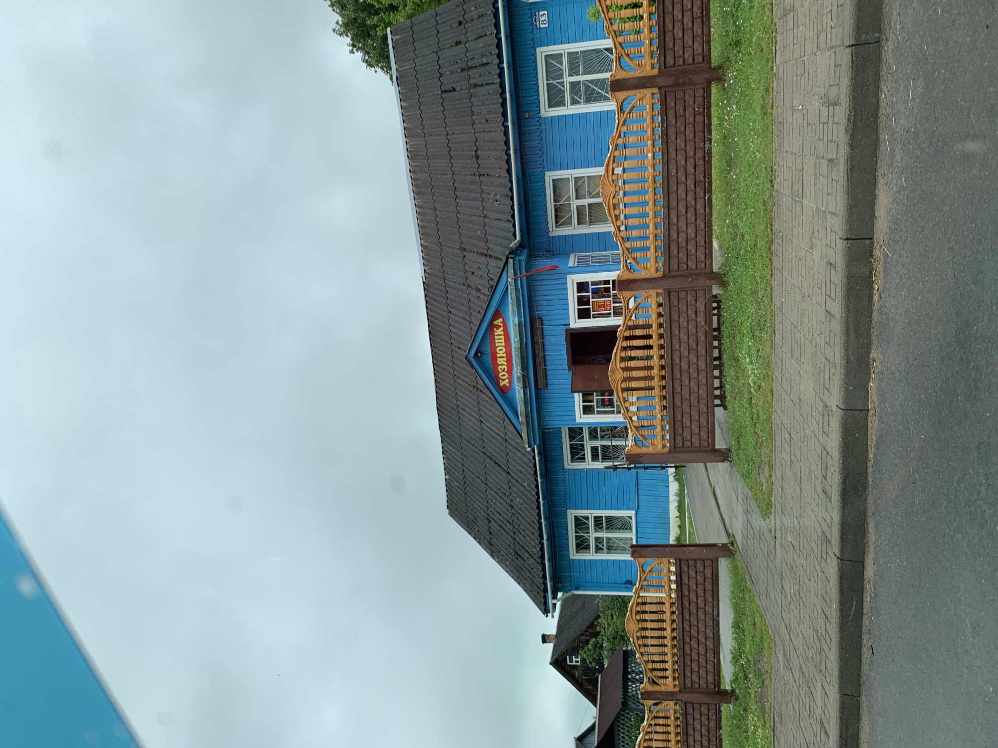 The image depicts a bright blue wooden building with a dark grey tiled roof, identified as "ХОЗЯЮШКА" (Khozyayushka), situated in the city of Ilyanskiy selskiy Sovet, Belarus. The building has several white-framed windows, some with decorative grates. A red flag is visible hanging from the building. In front of the structure, a light grey paved walkway leads to an entrance. This walkway is flanked by a decorative fence, with brown brick-like lower panels and light brown, ornate wooden slats above. A green grassy area with small white flowers is visible beyond the fence. The scene appears to be a commercial establishment, possibly a shop, given the sign and the operating hours of 9-18 displayed on a window. The sky is overcast and grey. The image is taken from a street level perspective, with a portion of an asphalt road visible in the foreground.