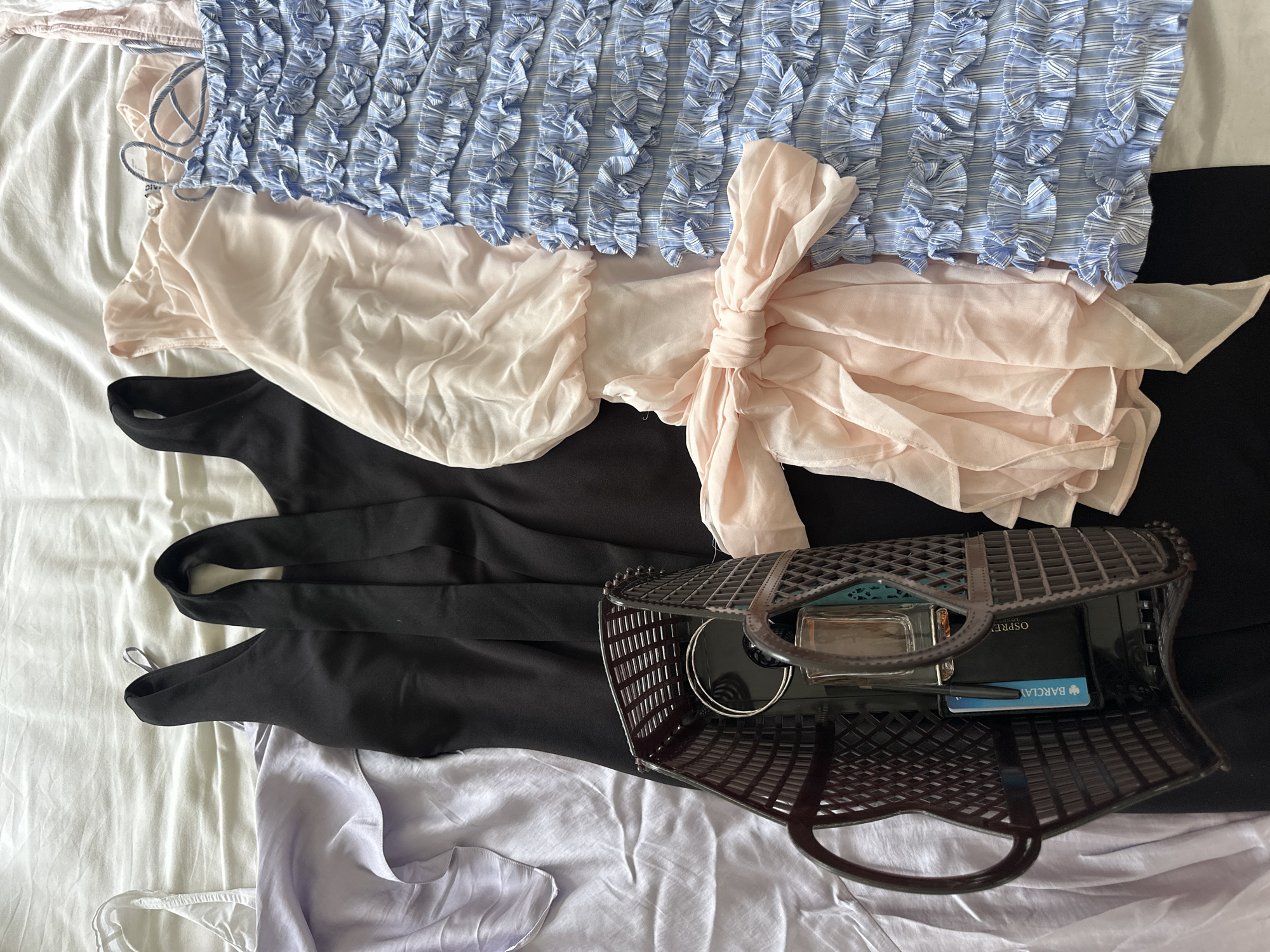 The image shows a pile of clothing and personal items on a white surface, likely a bedsheet. Clothing items include a light blue and white striped top, a light pink draped garment, a black dress, and a light purple satin-like item. On top of the black dress is a dark brown plastic basket-style bag containing items such as a wallet, a perfume bottle, a Barclay's bank card, and a silver ring. The location is not discernable from the image. Therefore, there is no indication of this image being taken in Tunbridge Wells, United Kingdom.