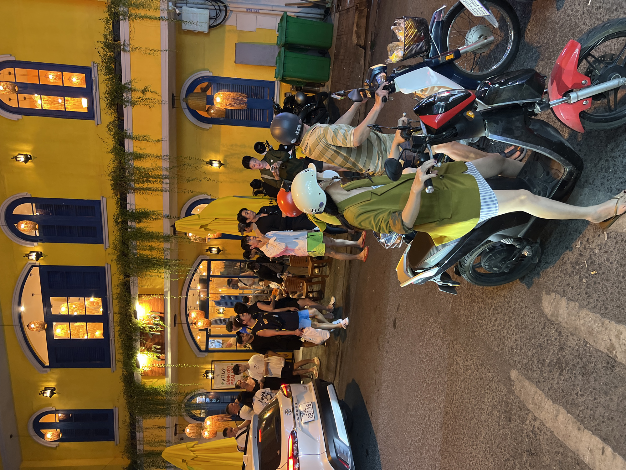 The image captures a bustling street scene at night in Nha Trang, Vietnam, likely near a restaurant. The prominent yellow facade of a building, illuminated by warm interior lights and decorative hanging lamps, dominates the background. Blue shutters are visible on arched windows. Green vines cascade down from above the windows, adding a touch of nature to the urban setting.
In the foreground, two people are seated on a black and red scooter. The person in front, wearing a green jacket and a white helmet, has their left hand on the handlebars. They are wearing a horizontally striped skirt. The person behind them, wearing a striped t-shirt, is also on the scooter. Next to them is a blue and white scooter, with a basket containing items.
A white car, a Toyota Cross, is parked on the left side of the street, facing away from the viewer. Its license plate reads "30L 097.18". Several groups of people are walking on the sidewalk and street, some appearing to be patrons of the establishment. There are also two green trash bins visible on the right side of the image. The asphalt street is lit by artificial lighting, creating reflections and shadows.