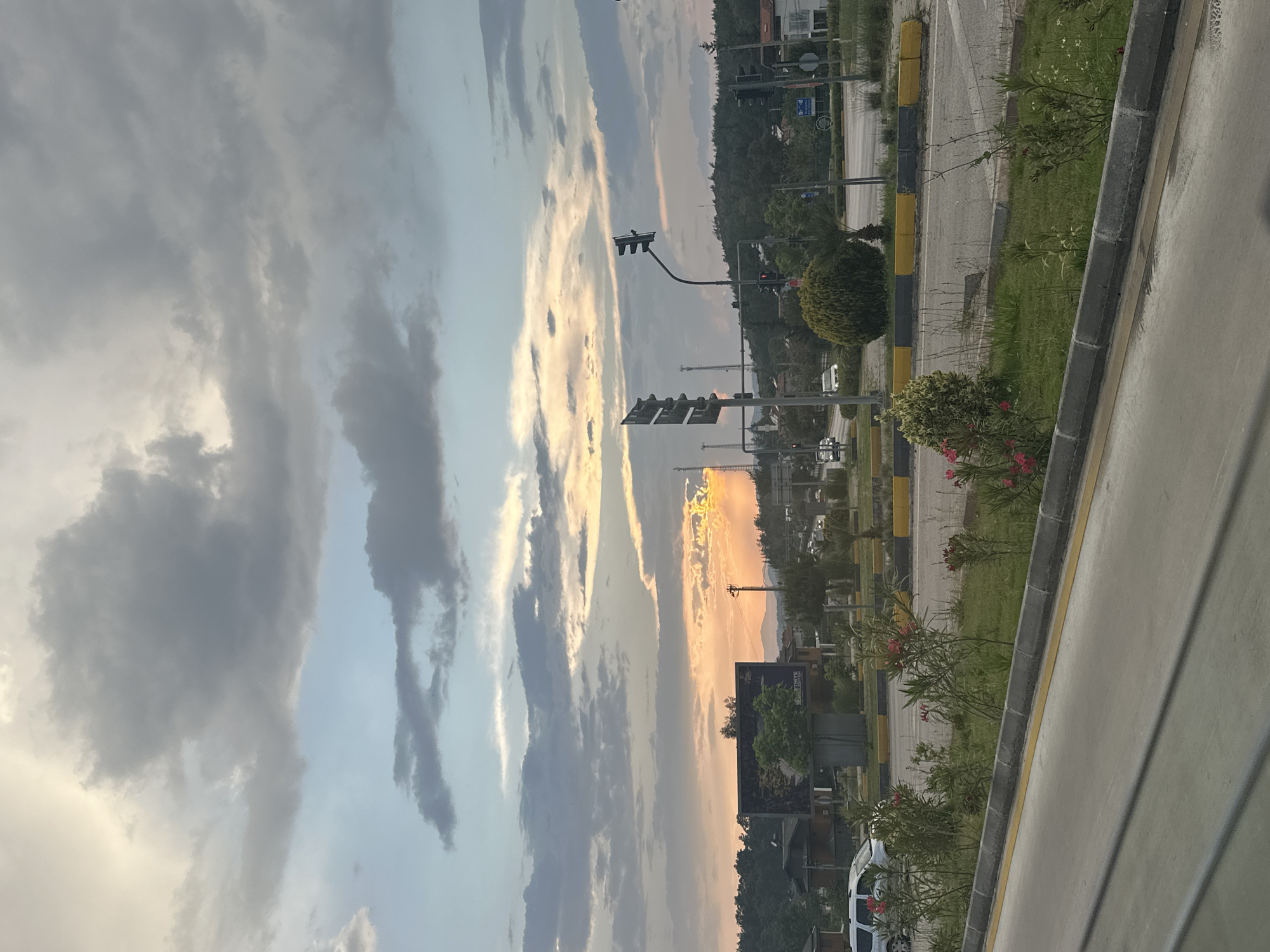 The photograph presents an urban landscape in Fethiye, Turkey, taken during twilight. The scene is oriented vertically with the sky dominating the left portion of the frame, filled with stratocumulus clouds illuminated by the setting sun. The clouds transition from pale gray to blue, and the setting sun casts a golden glow between the clouds. The right half features a roadside view of an urban area. The street has a curb that separates it from the green grass lawn. Bushes and other greenery run along the curb. Streetlights and traffic signals are visible. Beyond the street are buildings. A billboard sits along the roadside. The scene is static with no obvious human activity. The photograph gives the impression of a quiet scene.
