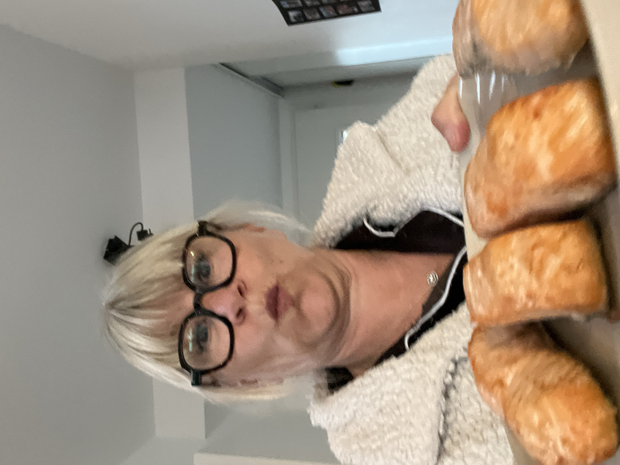 The image shows a woman wearing glasses and a light-colored, textured jacket. She is holding a container with several small, bread-like rolls inside. The background appears to be an interior space, possibly a home, with a hallway visible. The location in the city of Chambœuf, France is not verifiable from this image alone.