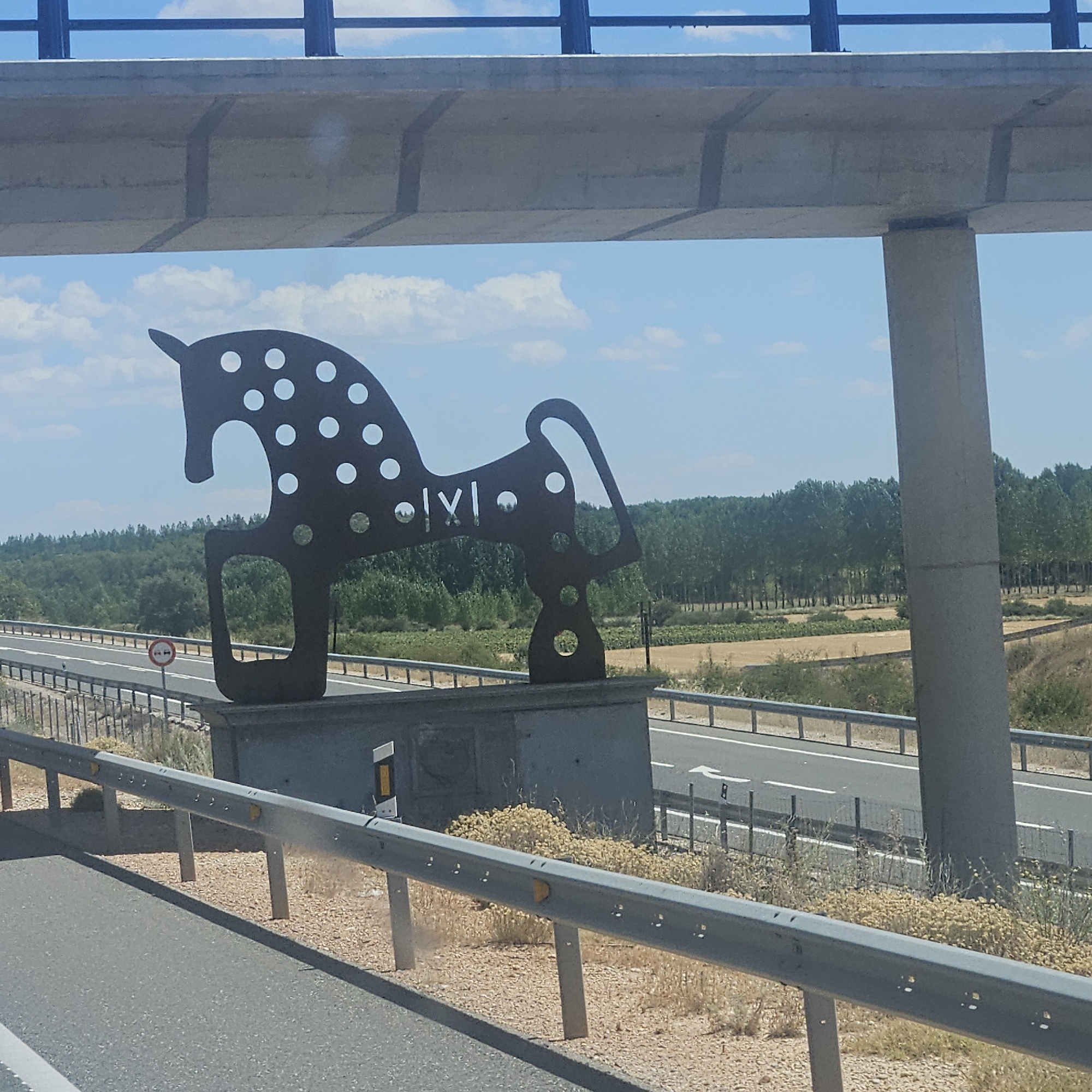 The image shows a roadside sculpture of a horse with white polka dots, positioned on a grey pedestal. The sculpture is dark metal and features cut-out circles on its body, with Roman numerals "IXI" on its flank. It is located near a highway with a concrete overpass and guardrails in the foreground. The background consists of a line of trees and a clear blue sky with scattered clouds. A road sign with red circles is visible on the left side of the highway. This sculpture is located in San Esteban de Gormaz, Spain, and is a landmark on the A-11 highway, specifically the Autovía del Duero.