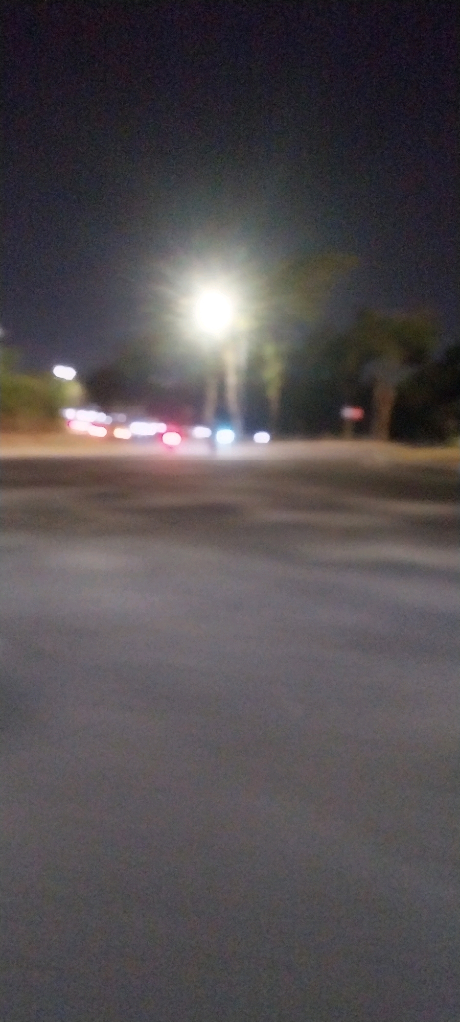 The image depicts a blurry night scene on a street, likely in Medang, Indonesia. A bright streetlamp illuminates the area, casting a glare. In the background, several cars with their headlights and taillights illuminated are visible on what appears to be a road or intersection, suggesting vehicle traffic. Trees and some indistinct structures can be seen behind the road. The foreground is dominated by a dark, asphalt-like surface, possibly a road or sidewalk. The overall impression is one of a moderately lit urban environment at night, with moving vehicles contributing to the bokeh effect. There are no discernible individuals or specific identifiable structures, given the blurriness of the image.