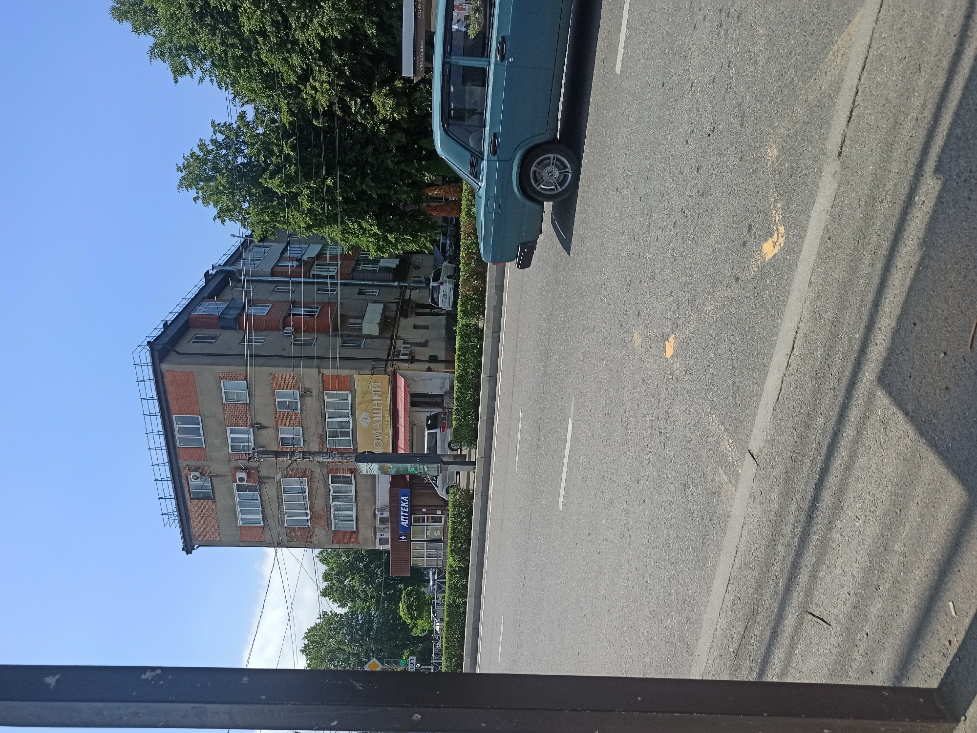 An eye-level outdoor medium shot captures a street scene in daylight, featuring a turquoise sedan driving away from the camera on a paved road. The car is in the right half of the frame, its rear end partially visible. The road has white lane markings and some yellow markings. A dark, vertical pole is positioned in the left foreground, casting a shadow. In the background, a multi-story brick and concrete building stands, with several windows and air conditioning units visible. A small storefront with blue signage saying "АПТЕКА" (Apteka, meaning Pharmacy) is located at the base of the building. Trees with lush green foliage line the street behind the building and across the road. The sky is clear and blue.