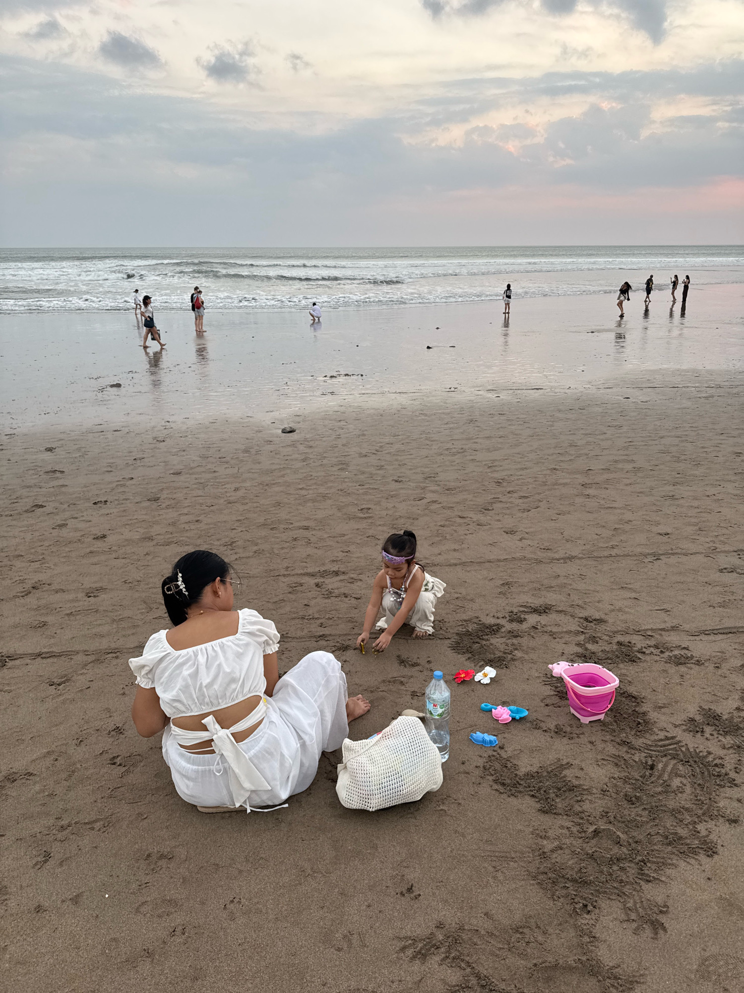 In Kuta, Indonesia, a mother and daughter are playing on a sandy beach. The mother, wearing a white backless top and white pants, sits with her back to the camera, facing the ocean. Her daughter, dressed in a light-colored outfit, squats in the sand in front of her, playing with toys. Scattered around them are a water bottle, a pink pig-shaped bucket, a blue shovel, and two small decorative flowers. The ocean is visible in the background with gentle waves rolling onto the shore. Several other people are present on the beach, some walking near the water and others further away. The sky is overcast with muted colors, suggesting it is either early morning or late afternoon. The sand is damp and textured with footprints and the marks of the tide.