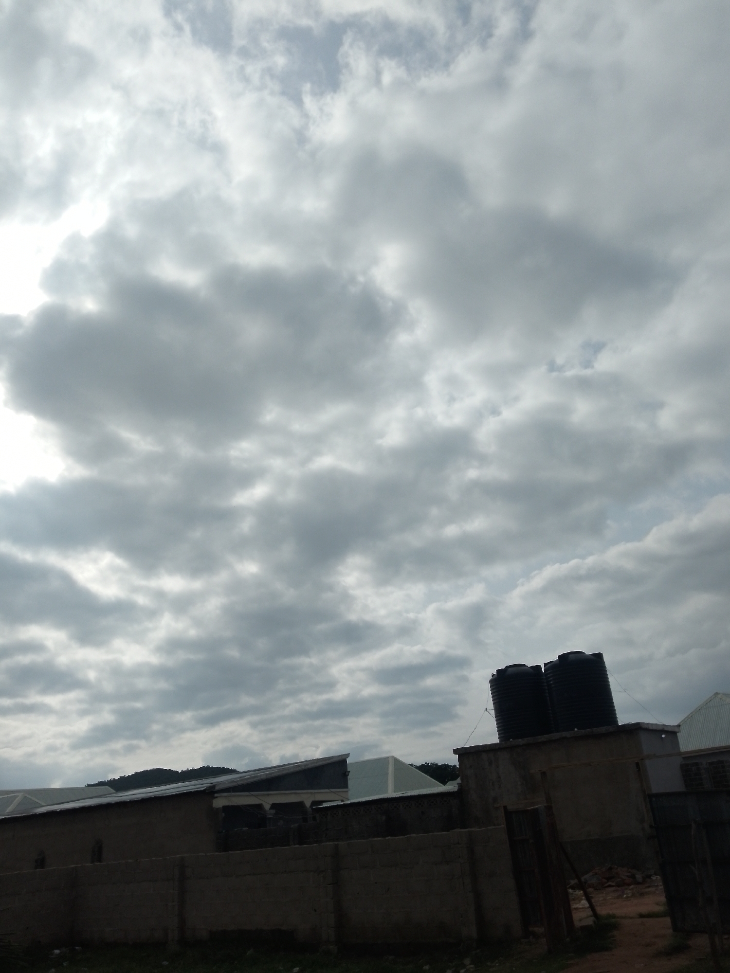 This is a wide-angle, eye-level shot of a cloudy sky above a low-rise building in Serti, Nigeria. The sky is dominated by heavy, grey clouds with patches of lighter sky visible. In the foreground, the top of a brick wall runs horizontally across the frame. Behind the wall, a building with a sloping roof is visible, topped with two large black water tanks. Another building, partially obscured, is to the right of the water tanks. The overall lighting suggests an overcast day.
