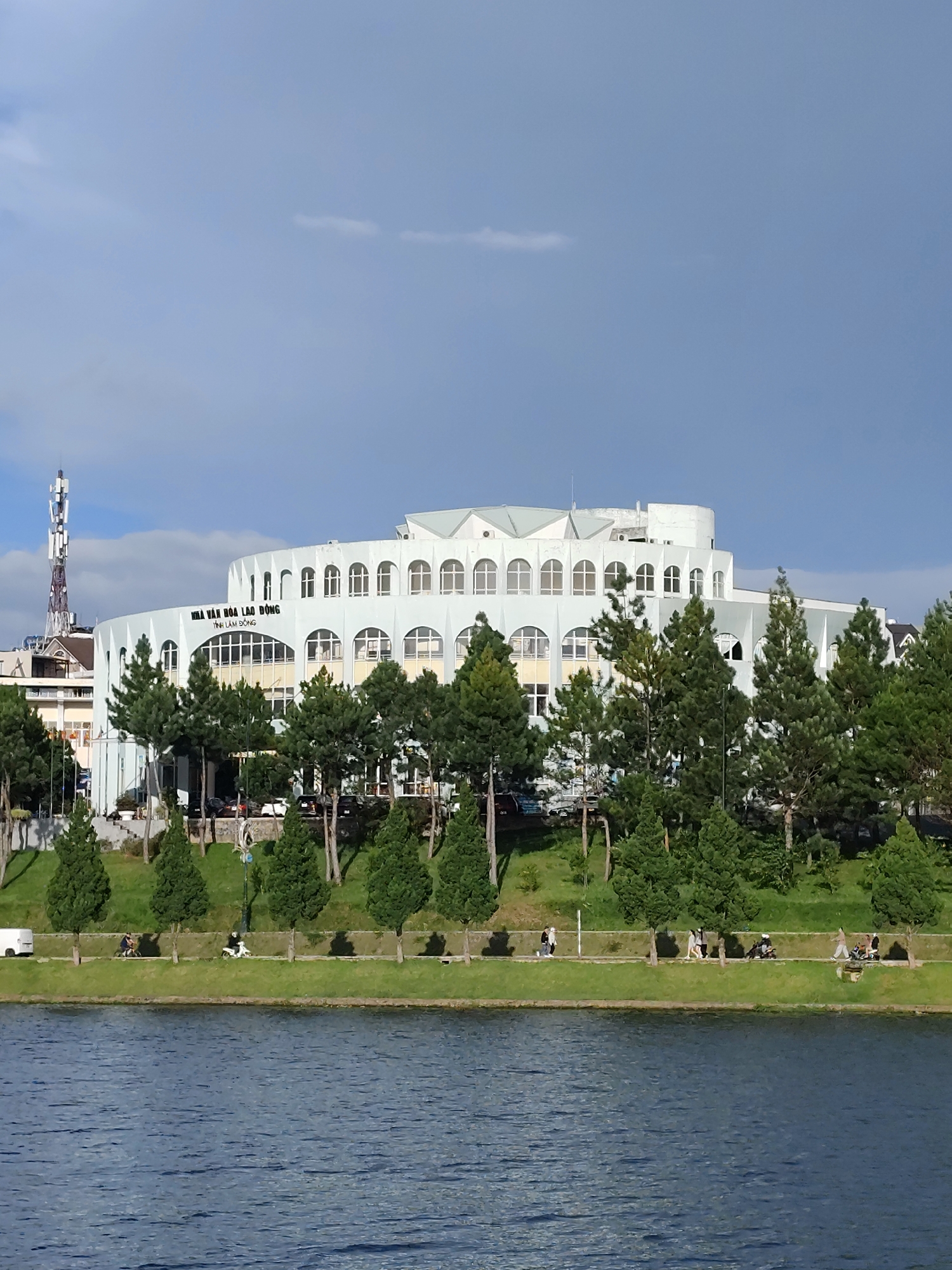 The image shows a large, modern, multi-story building with a curved facade, featuring arched windows. The building is situated in Xuân Hương Lake, Đà Lạt, Vietnam. In front of the building, there are many pine trees. The lake is in the foreground, and a grassy embankment with a walking path runs along its edge. Several people are visible on the path, some walking and others riding motorcycles. A white van is parked on the left side of the path. A tall cell tower is located to the left of the main building. The sky is blue with some scattered clouds.