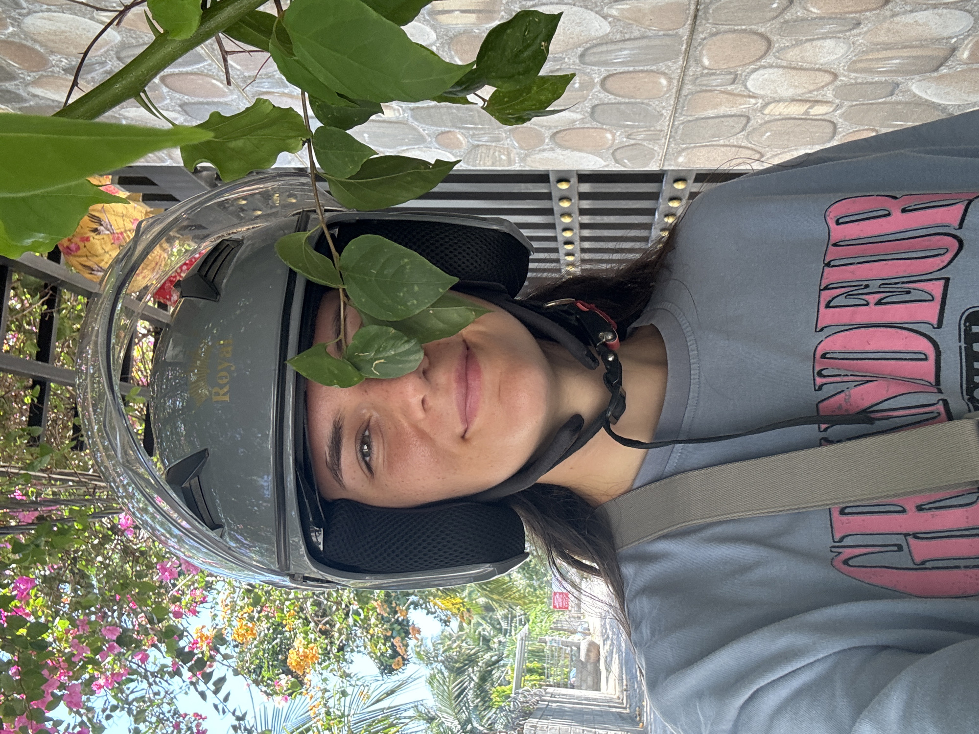 A woman is photographed wearing a gray helmet labeled "Royal," with green leaves from a nearby plant extending across the helmet and slightly obscuring part of her face. She has fair skin, dark hair, and is wearing a gray t-shirt with a pink design. A light gray strap is visible diagonally across her shoulder. The background features flowering bushes with pink and orange blossoms, palm trees, and a pathway. The ground is paved with gray cobblestones. The photograph appears to be taken at a roadside in Mũi Né, Vietnam.