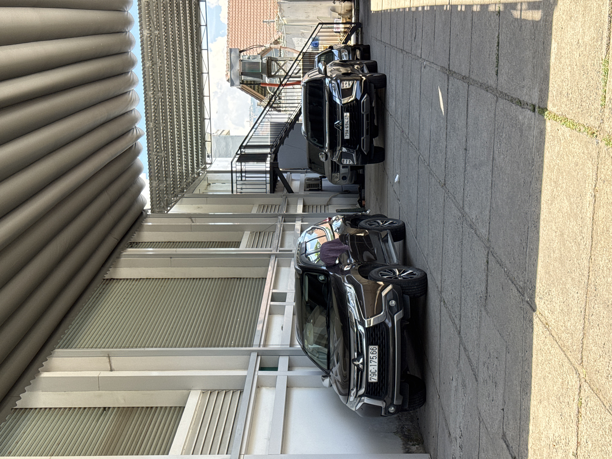 The image shows a row of parked cars in an alleyway. The cars are all dark-colored SUVs, and appear to be from the same model or make. The alleyway is paved with concrete, and there are buildings on either side. One building has a corrugated metal roof and metal shutters. There is a set of stairs leading up to a balcony or upper floor. The lighting suggests it is daytime, with sunlight casting shadows on the ground. The image appears to be taken from a slightly elevated position. The visible license plate on one of the cars reads "79C-175.66". The overall scene depicts a typical urban or suburban parking area.
