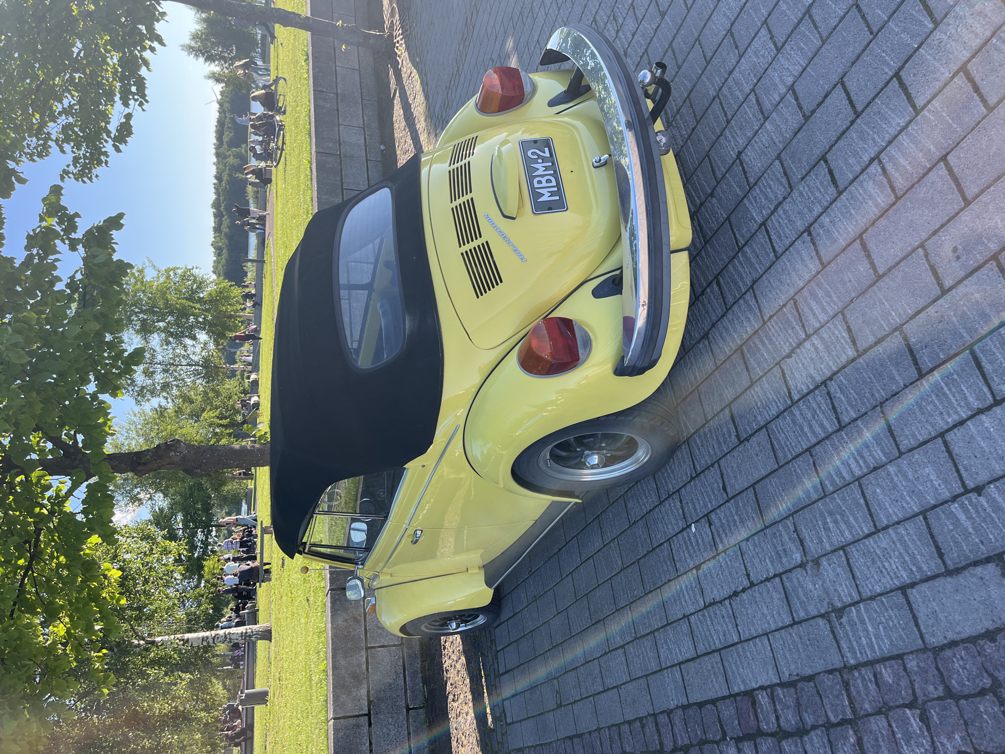 A yellow Volkswagen Beetle convertible is parked on a paved walkway by a stone wall. The car's black soft top is up. In the background, beyond the wall, is a grassy area with trees and a body of water. Several people are scattered across the grass, some sitting and others standing, facing a lake or river. The scene appears to be set in a park or recreational area in Oulu, Finland, possibly during a summer day, given the clear sky and the presence of people outdoors. The license plate on the car reads "MBM-2".