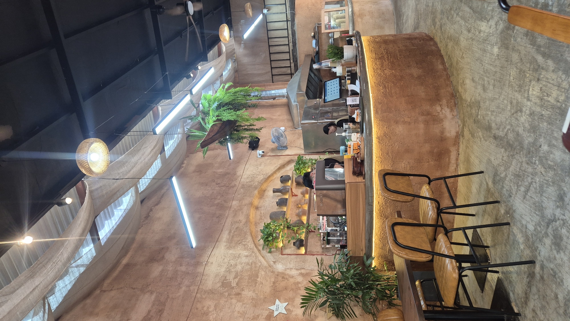 This image depicts the interior of a coffee shop located in Da Nang, Vietnam. The coffee shop has a rustic and bohemian aesthetic. The walls are textured in a warm, earthy tone, and draped fabric hangs from the ceiling, creating a soft, diffused lighting effect.
In the foreground, two brown leather chairs with black metal frames are positioned at a dark wooden table. The floor is concrete.
The central focus is a curved counter, also with a textured, earthy finish, illuminated by a warm yellow LED strip light along its base. Behind the counter, a coffee machine and various barista equipment are visible. Two baristas are present: one is partially visible behind the coffee machine, and another is serving a customer at the counter. A computer monitor displaying what appears to be a menu or order system is on the counter.
Shelving with decorative vases and plants is mounted on the arched wall behind the counter. Various plants are placed throughout the shop, including a large potted plant to the left and hanging plants suspended from the ceiling, one of which is in a unique boat-like planter.
Overhead, a large, woven pendant light hangs, and fluorescent strip lights are also installed. A ceiling fan is visible in the background. The overall ambiance suggests a casual and inviting coffee shop environment.