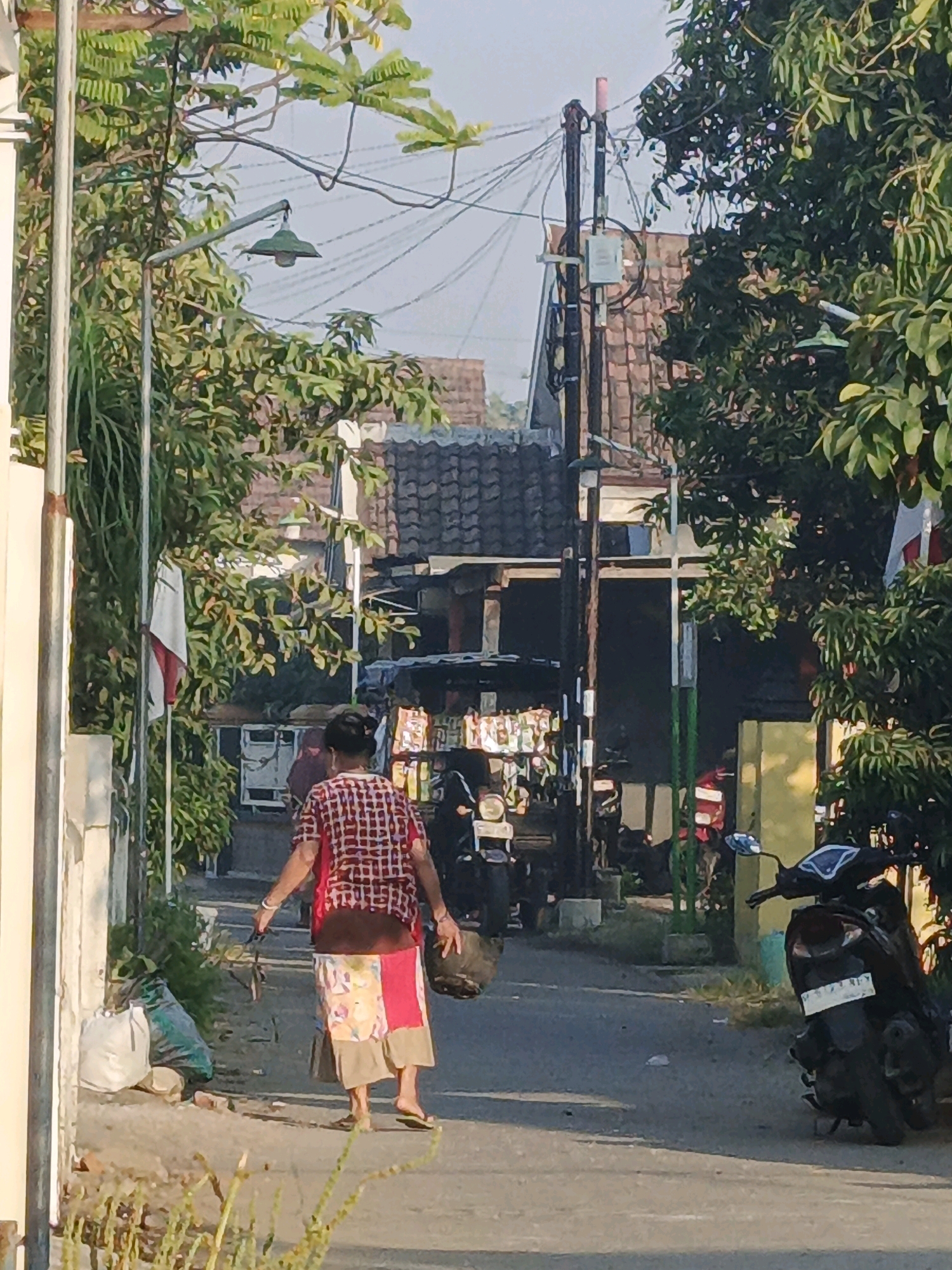 A full-shot, eye-level, outdoor medium shot captures an individual walking away from the camera on a paved street lined with trees and buildings. The person, with dark hair pulled back, is wearing a red and white checkered short-sleeved shirt and a colorful, patchwork skirt. They are carrying a dark bag in their right hand and appear to be holding something in their left hand, possibly a leash. To the left of the street, a white wall is visible, with a large white bag and a green bag on the ground in front of it. A light pole with a hanging lamp is on the left. Further down the street, there are parked motorcycles and a structure with a tiled roof. The scene appears to be in a residential area in Ketegan, Indonesia, indicated by a flag on the left and the architectural style of the buildings. The lighting suggests it is daytime, with sunlight casting shadows.