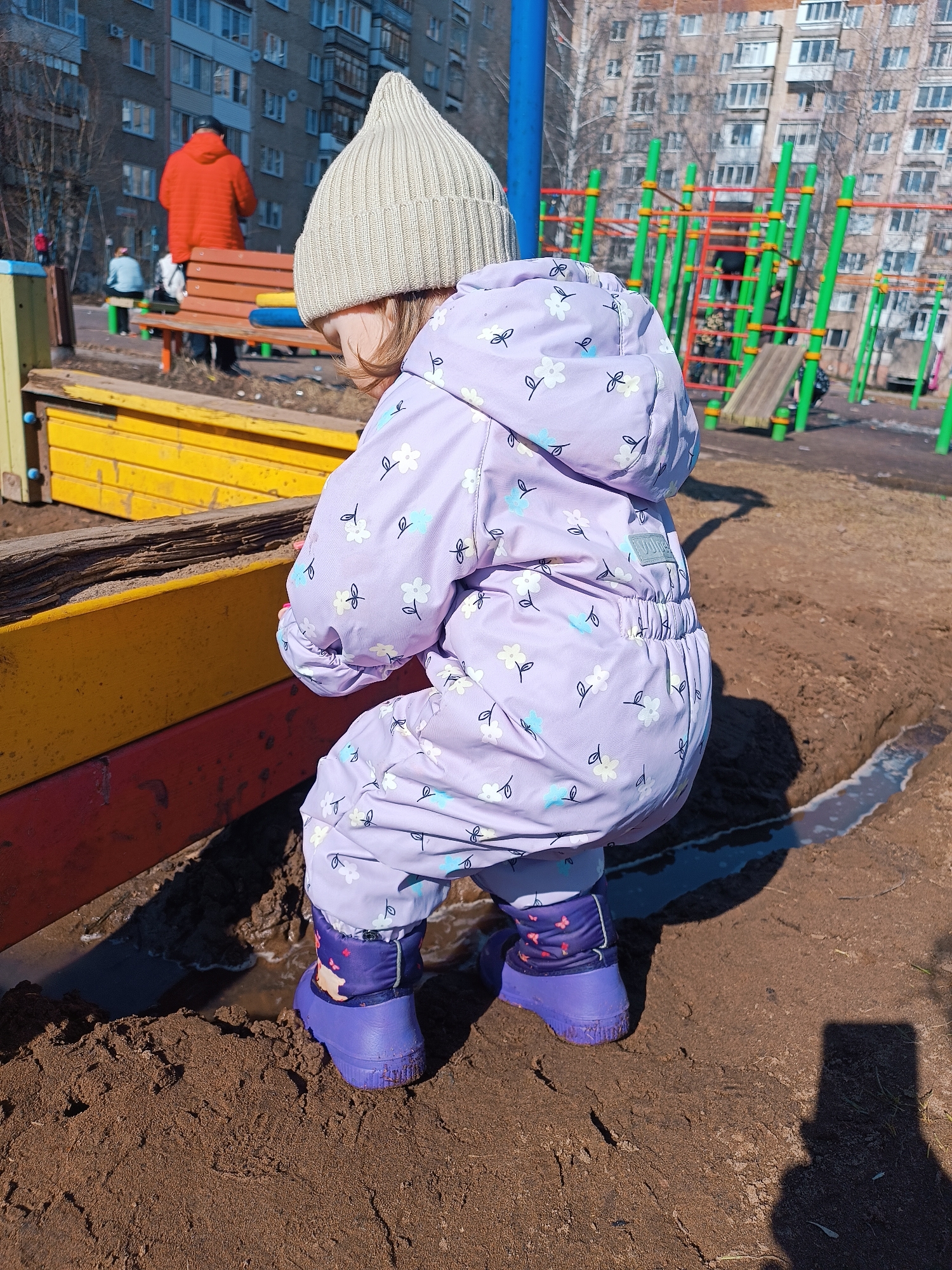 In Izhevsk, Russia, a young child in a lavender snowsuit with a white floral pattern and purple boots is playing in a muddy area beside a colorful sandbox. The child wears a tan knit hat. In the background are apartment buildings, playground equipment, and figures of people on benches. The ground is a mix of mud and dirt. The sandbox is painted yellow, red, and blue. A shadow, possibly of a person taking the photo with a phone, is visible on the ground to the right.
