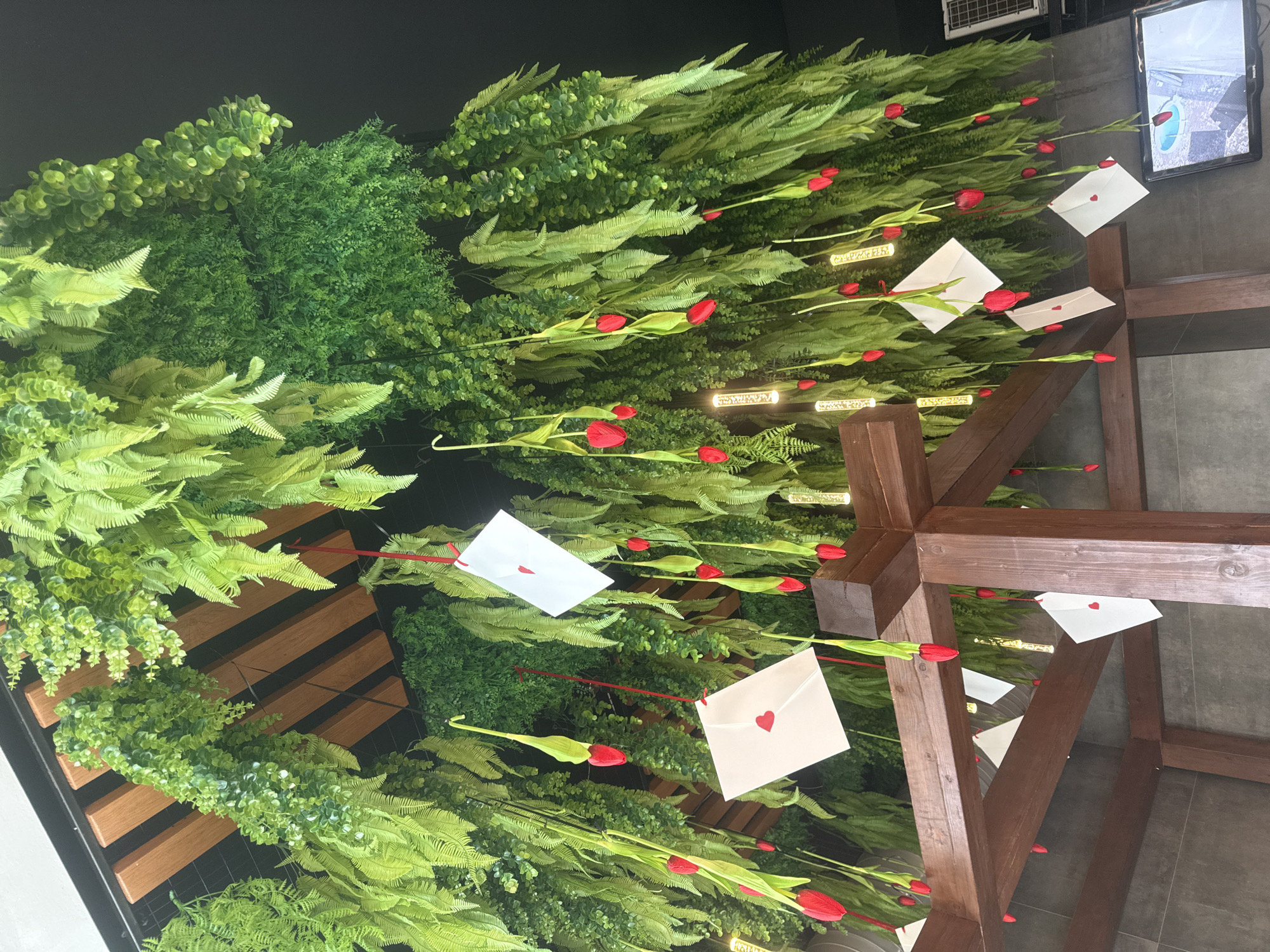 A view looking up at a ceiling installation, possibly indoors in Glazov, Russia. The installation features artificial greenery of various types, including fern-like plants and smaller leafy bushes. Red tulip buds are interspersed among the greenery, attached to thin green stems. White envelopes, each decorated with a small red heart, are suspended among the plants. A dark brown wooden structure, possibly a decorative trellis or frame, intersects the display. In the background, there is a dark wall with what appears to be a television screen. The overall effect is a blend of natural and artificial elements, creating a romantic or decorative atmosphere.