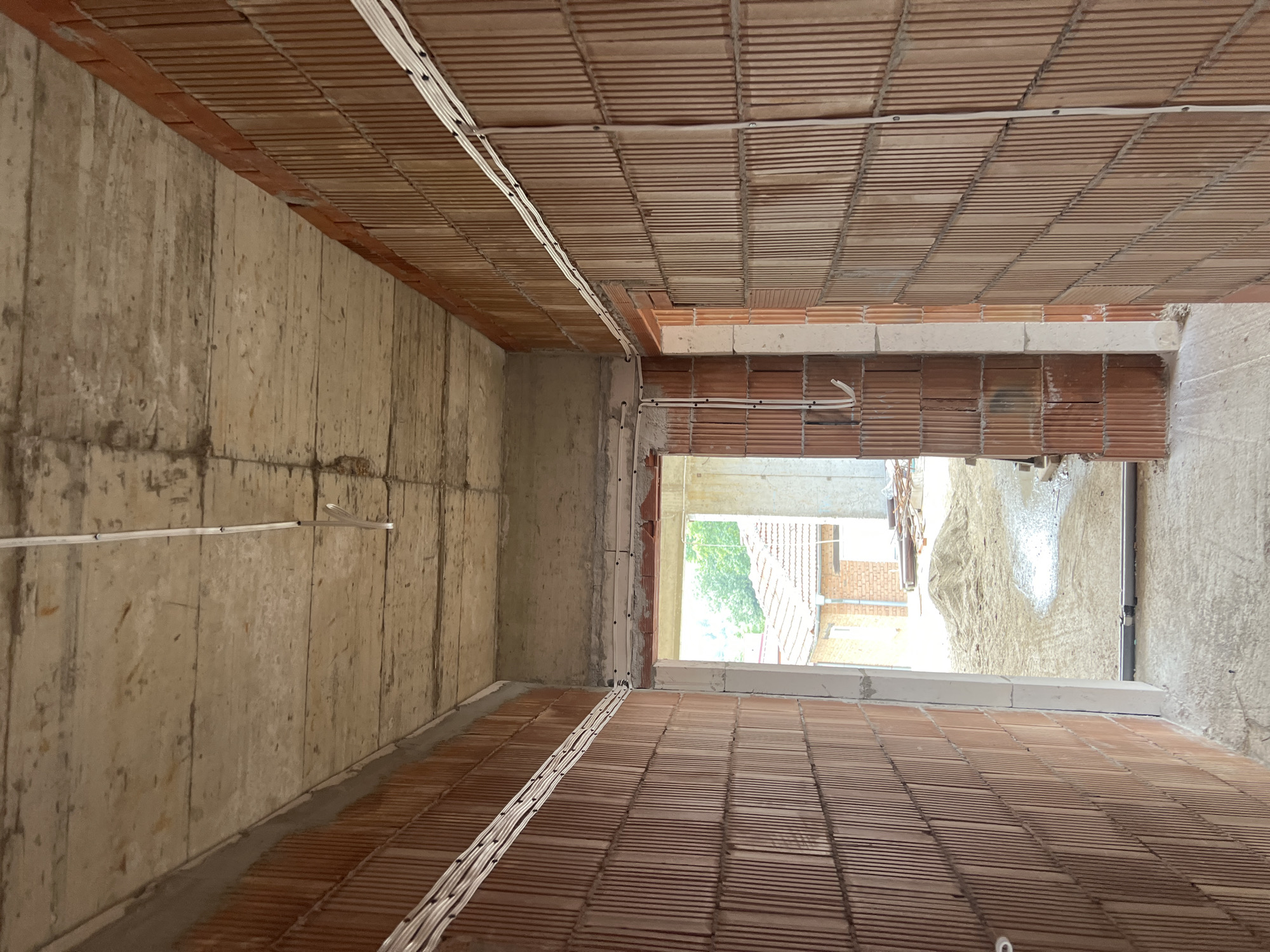 This image captures the interior of an unfinished building, looking down a corridor or room under construction. The perspective is a downward vertical shot.
The left wall is made of rough, unpainted concrete, exhibiting visible vertical lines likely from wooden formwork. The right wall and the ceiling are constructed from exposed, hollow, reddish-brown clay blocks or tiles, arranged in parallel rows, creating a textured, ribbed surface. White electrical cables, bundled together and secured with clips, are visibly routed across the ceiling and along the upper sections of the walls, indicating the early stages of electrical installation. Some of these cables also extend along the floor, which appears to be constructed from similar reddish-brown clay blocks.
At the far end of the space, a large opening, possibly a future doorway or window, provides a view to the outside. Bright daylight streams through this opening, illuminating distant green trees or bushes, a building with a red tiled roof, and a prominent pile of sand or dirt directly outside the opening. A concrete lintel or beam is visible above this opening.
The scene suggests an active construction site, characterized by raw building materials and the exposed roughing-in of utilities. No people are visible. The bright light from the exterior indicates it is daytime. The construction style and materials are typical for a building in Topolovo, Bulgaria. There is no visible text in the image.