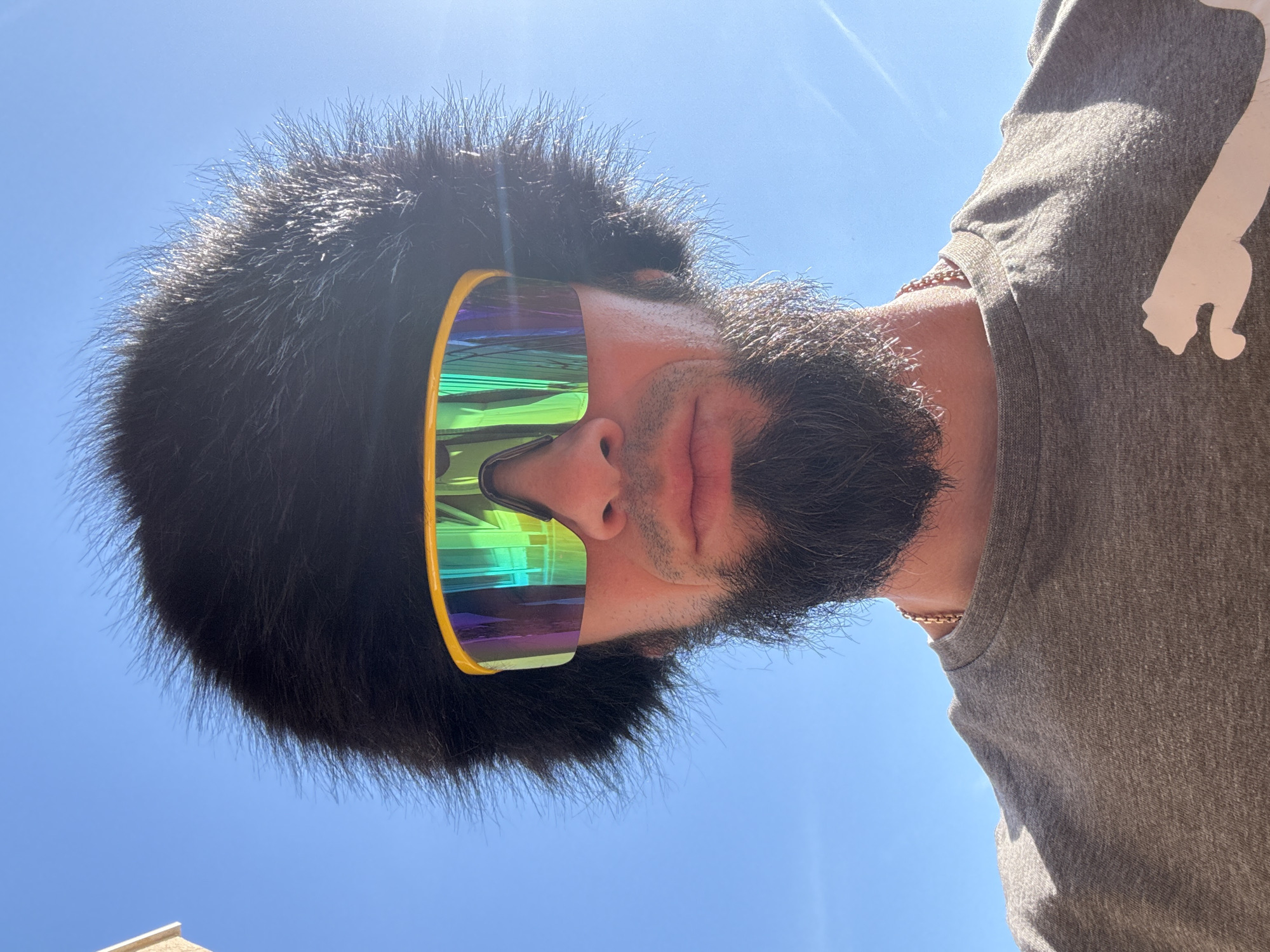 A low-angle, close-up shot shows a man with a dark beard and hair wearing large, iridescent sports sunglasses with yellow frames. He is wearing a gray t-shirt with a white design on the right shoulder. A gold chain is visible around his neck. The background is a clear blue sky, suggesting a sunny day. The image was taken in an outdoor environment. There is no indication of Šušanj, Montenegro from the image.