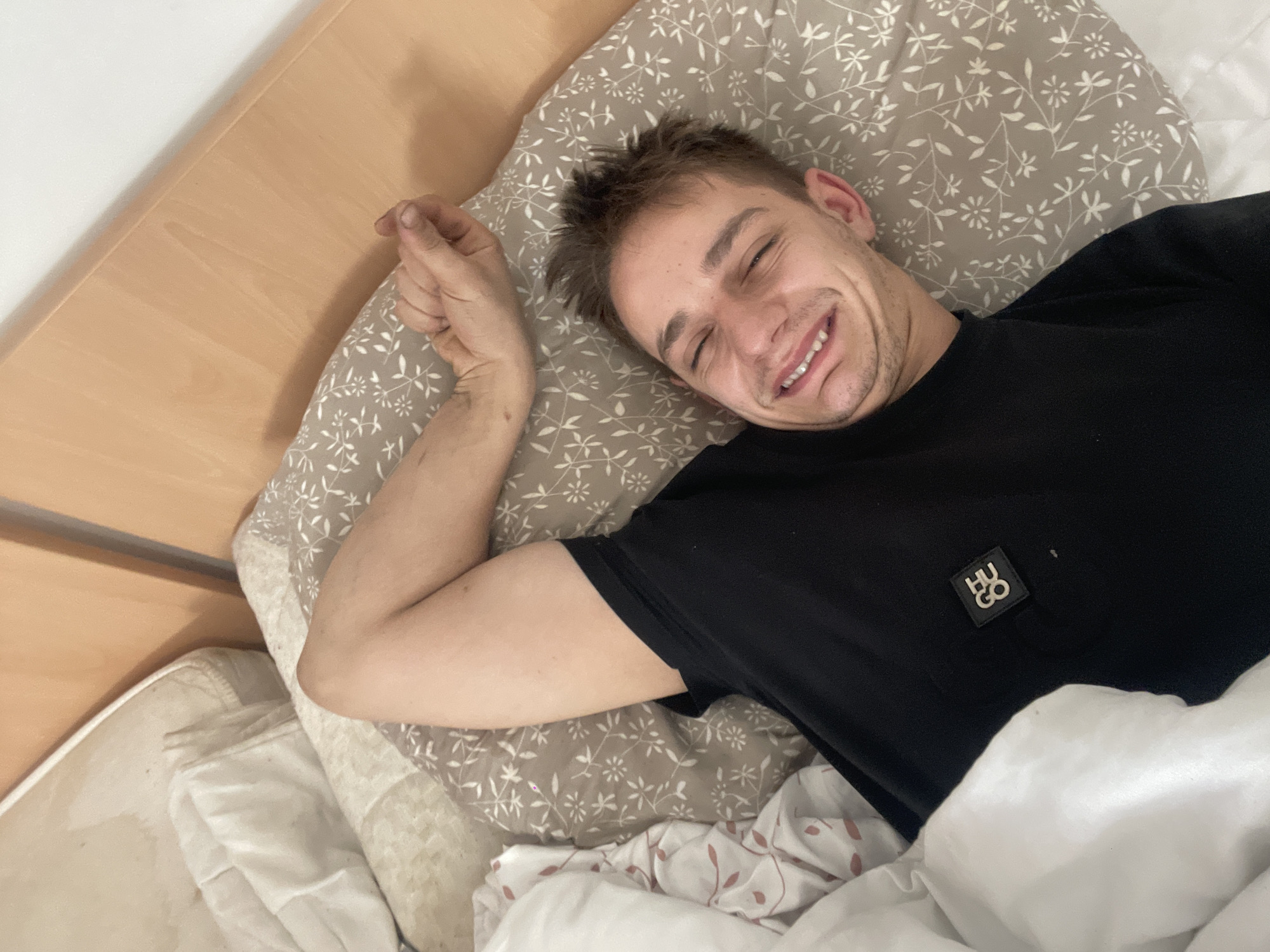 A young Caucasian male is lying in bed, facing the camera with a wide, open-mouthed smile. He has short, light brown hair and is wearing a black t-shirt with a small "HUGO" logo on the left chest. His right arm is bent at the elbow and resting behind his head, with his hand near the pillow. The background consists of a bed with a light brown, floral-patterned pillow and white bedding. Part of a wooden headboard is visible on the right side of the image. The lighting suggests an indoor setting, possibly a bedroom.