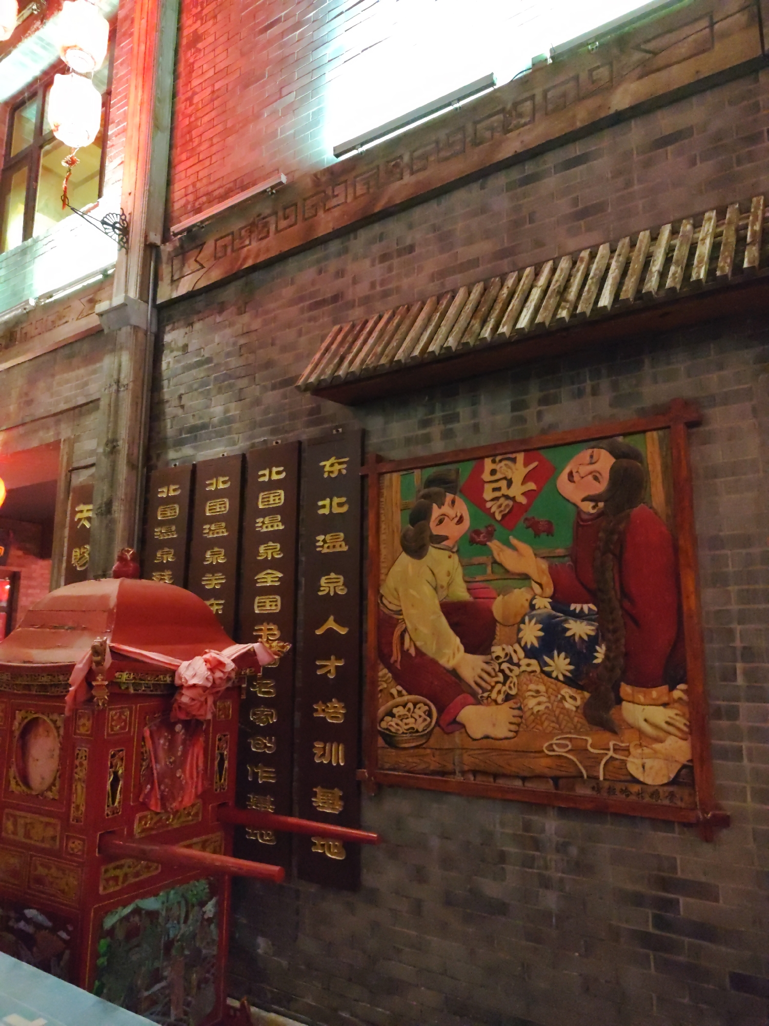 The image captures a street scene in Lindian, China, with a focus on traditional Chinese culture. The foreground features a vibrantly red, ornate palanquin, possibly used in processions or ceremonies. To the right of the palanquin, a large, wooden relief carving adorns the brick wall, depicting two figures in traditional attire. The carving is colorful and detailed, illustrating an interaction between two people. Above the carving, a wooden overhang with slatted tiles runs along the brick wall. Further to the right, vertical wooden plaques with golden Chinese characters are mounted on the wall, likely representing inscriptions or signage. The background consists of a brick building with visible windows and a warm, red brick facade. Chinese lanterns, illuminated and hanging, are visible on the left side of the image, contributing to the festive or cultural atmosphere. The overall scene suggests a location within a historic or culturally significant area of Lindian, China, emphasizing traditional aesthetics and artistic expression.