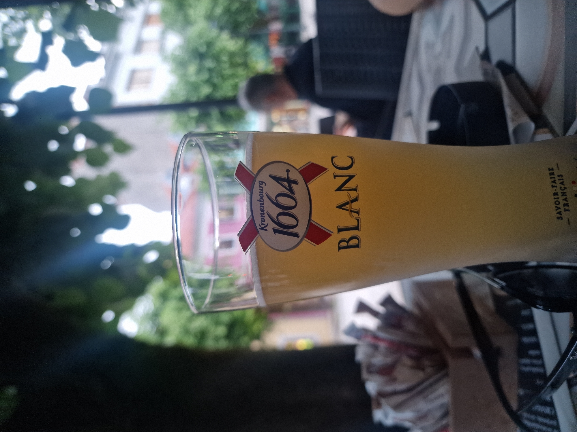 A close-up, slightly angled shot focuses on a tall, clear glass filled with pale yellow beer, topped with a white foam head. The glass is positioned on a patterned surface, likely a table. The label on the glass reads "Kronenbourg 1664 BLANC" with French text at the bottom that is partially obscured but appears to say "SAVOIR-FAIRE FRANÇAIS".
In the background, out of focus, a person with gray hair is seated at a table, facing away from the camera, with their hands on the table. Behind them, the scene appears to be an outdoor setting, possibly a cafe or restaurant. There are trees with dappled sunlight filtering through their leaves, and buildings with windows are visible in the distance. The overall atmosphere suggests a relaxed, casual outdoor environment. The city of Cetinje, Montenegro, is indicated as the location.
