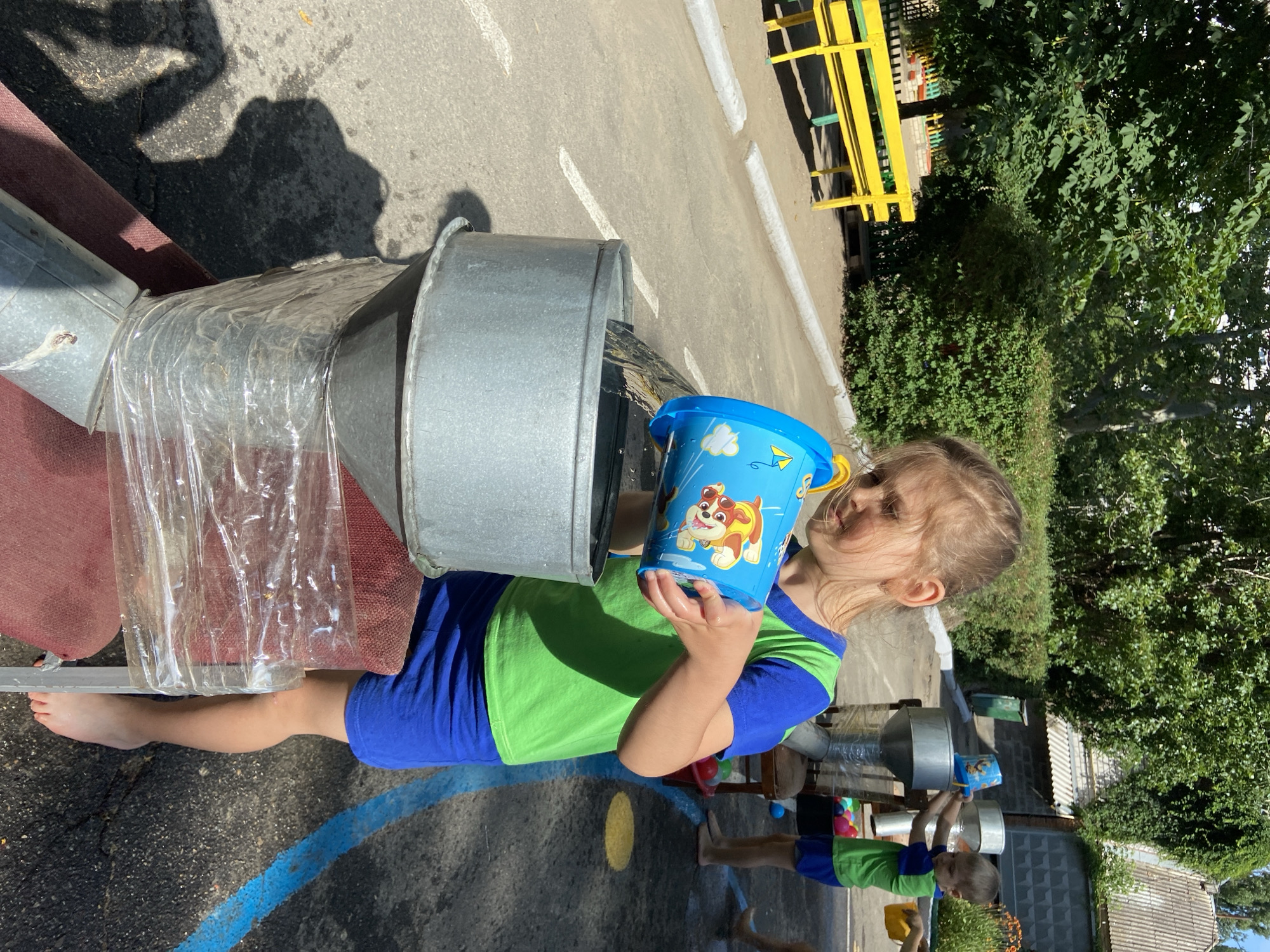 In this image, a young girl with blonde hair is playing outside on a sunny day. She is wearing a green and blue t-shirt and blue shorts. She is holding a blue bucket decorated with characters from Paw Patrol, seemingly pouring water into a large metal contraption that appears to be a water play structure. This structure is made of metal tubes and a clear plastic material, with water flowing through it. The girl's face shows concentration as she engages in her activity.
Behind her, another child, a boy also wearing a green and blue t-shirt and blue shorts, is visible. He is also holding a yellow bucket. They appear to be in an outdoor play area, likely a playground or a schoolyard, given the presence of a yellow bench and lush green trees in the background. The ground is asphalt, marked with white lines and a blue line. The lighting suggests it is daytime, with shadows cast on the ground, indicating a sunny environment. The overall atmosphere seems to be one of summer fun and outdoor recreation.