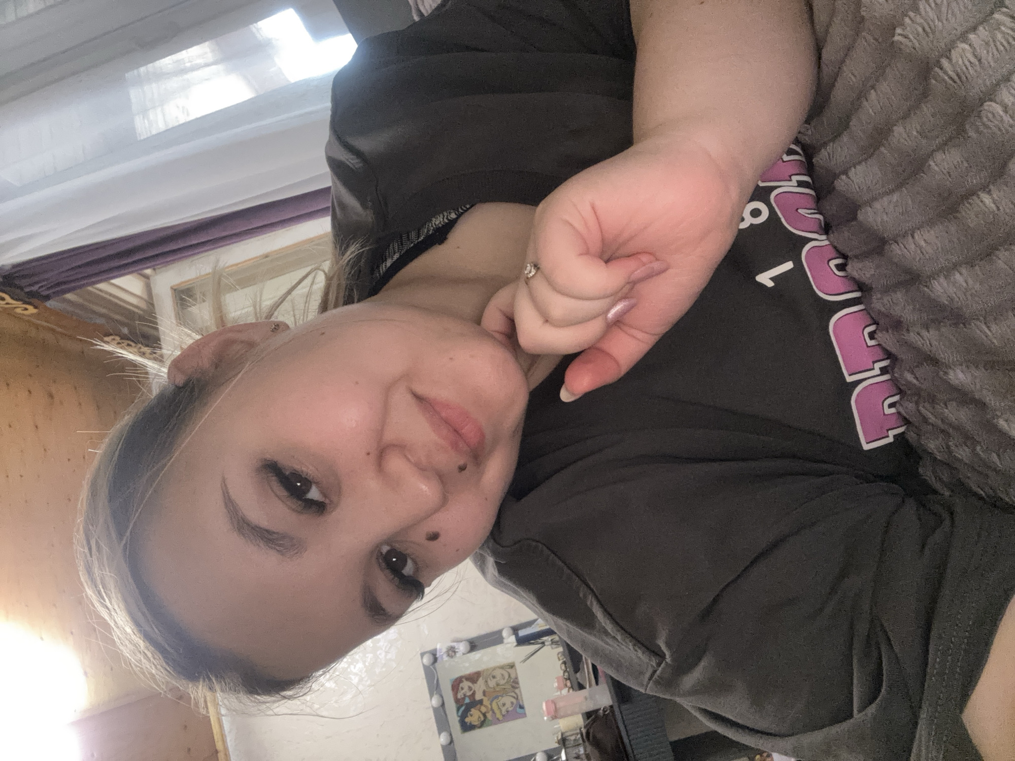 A young woman with light brown hair and dark eyelashes is lying down and looking at the camera. She is wearing a dark grey t-shirt with pink and white lettering on it. Her left hand, adorned with a ring and light pink nail polish, rests on her chest. The background shows a ceiling made of wooden planks, a window with purple and white curtains, and a vanity mirror with framed pictures of Disney characters. The woman appears to be in a bedroom. The image does not provide any specific information about the location in the city of Большой Букор, Russia.