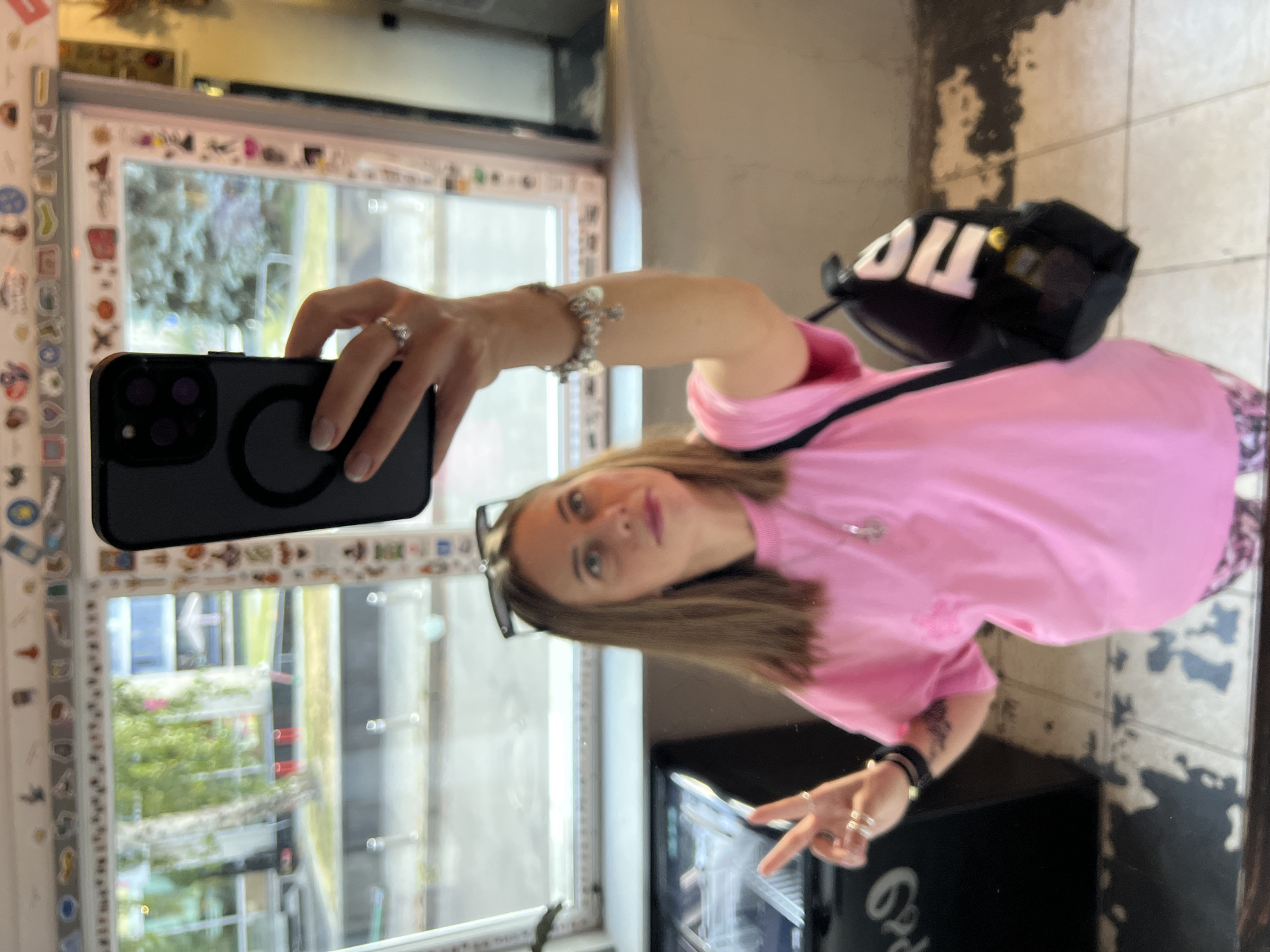 In Kyiv, Ukraine, a young woman is captured in a mirror selfie. She is wearing a bright pink t-shirt and patterned leggings. She has a backpack slung over her shoulder. Her hand holds a smartphone, taking the picture. The background suggests an indoor setting with a window overlooking an outdoor scene, possibly a street or park with trees. The window frame is decorated with various stickers.