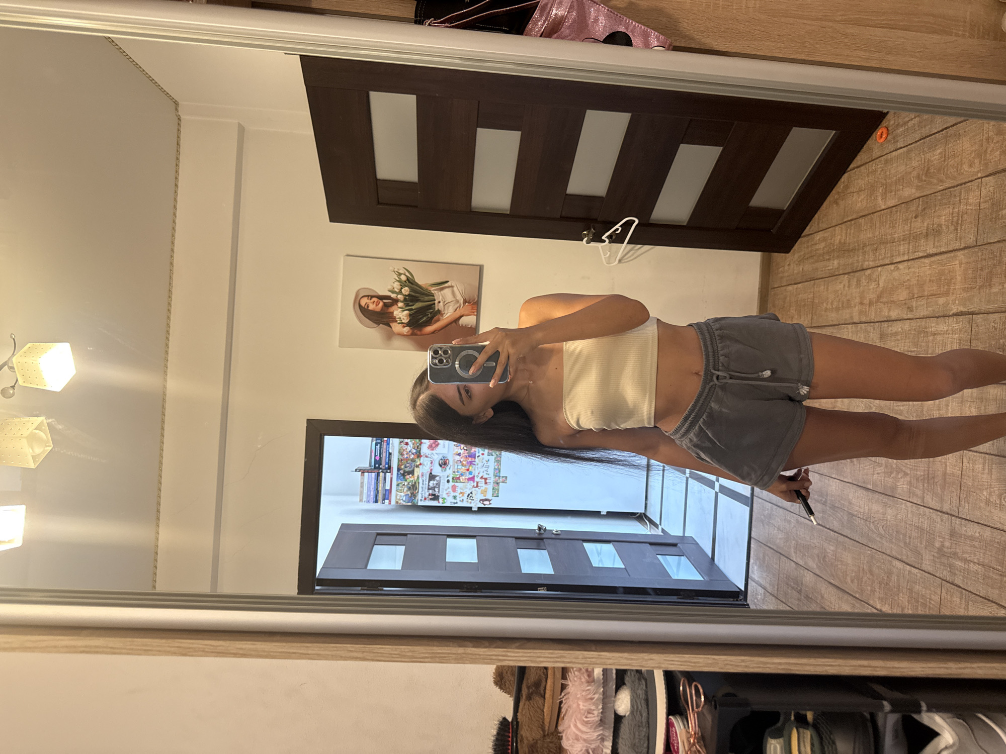 A young woman, appearing to be of Eastern European descent, stands in front of a large mirror, taking a selfie with her phone. She is wearing a white tube top and grey shorts. Her long, dark hair falls past her shoulders. In the background, an open door reveals a room with shelves of books and decorations. Another door, made of dark wood with glass panes, is visible to her right. The floor is made of wood planks. A framed picture of a woman holding a bouquet of tulips hangs on the wall. The setting appears to be a residential interior, likely an apartment or house. There are no specific elements in the image that would indicate the location as Chișinău, Moldova.