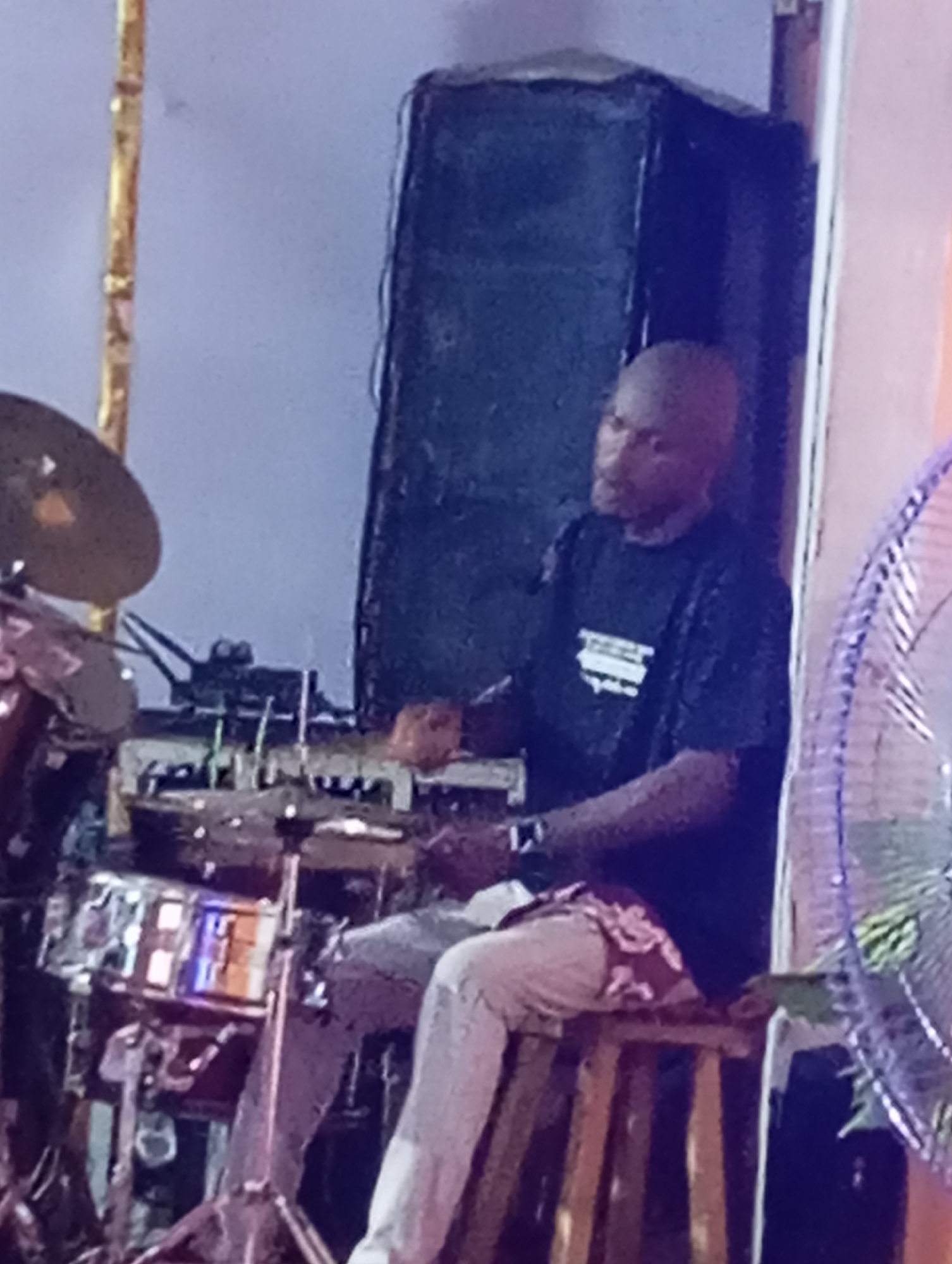 A man is playing the drums in what appears to be a performance venue. The drummer, who has a shaved head and is wearing a dark t-shirt and light-colored pants, sits on a wooden stool behind a drum kit. The kit includes cymbals, drums, and electronic equipment. Behind the drummer is a large speaker. A fan is visible to the right. The photograph was taken in Markurdi, Nigeria.