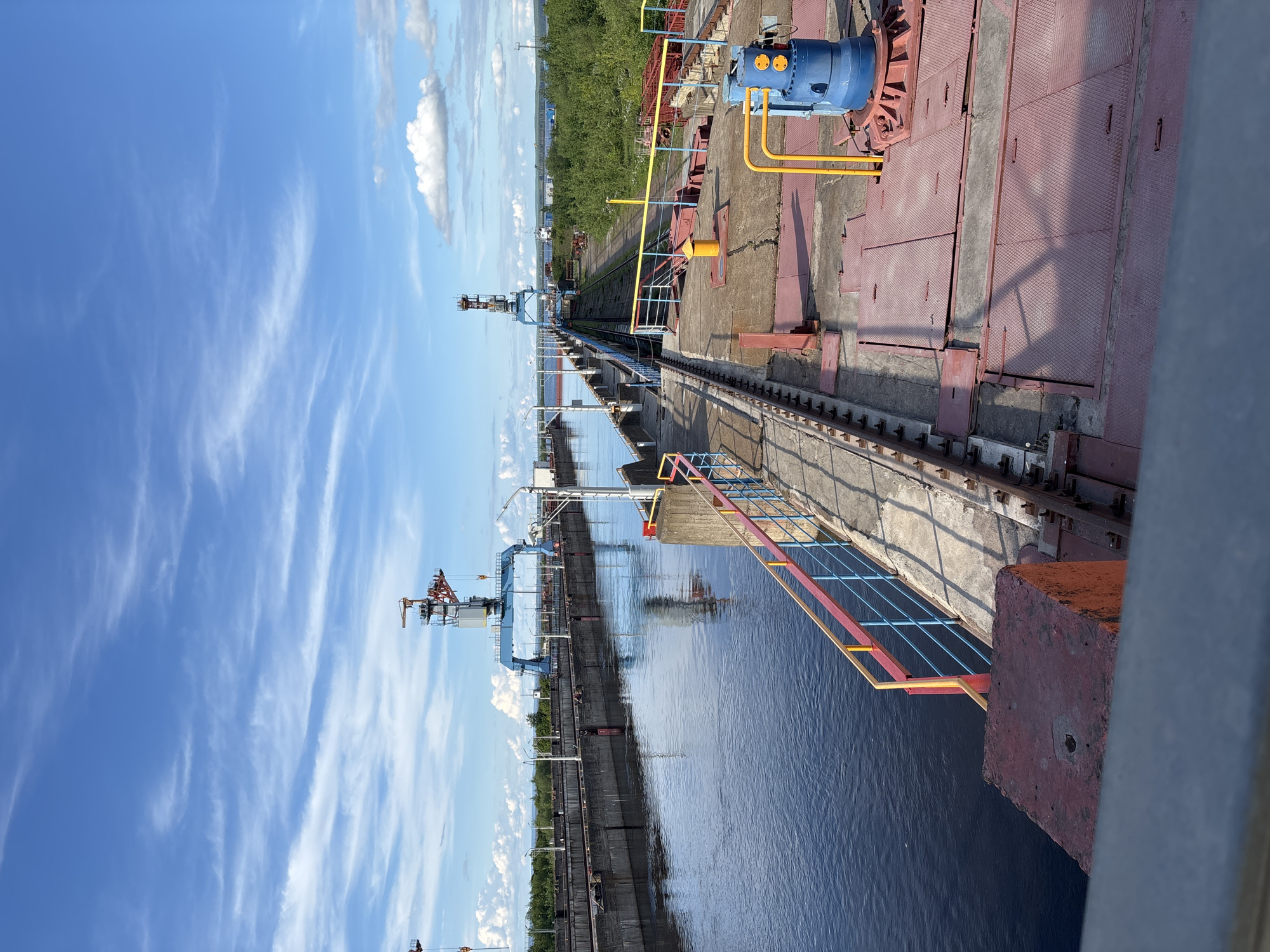 The image shows a section of the Volga-Kama Cascade hydroelectric station in Chaikovsky, Russia. A long concrete structure with metal rails runs along the water's edge. On the right, a red metal surface with a blue mechanical component is visible. The water reflects the blue sky and white clouds. Along the horizon, there are trees and industrial structures. The image appears to be taken from an elevated position on the hydroelectric station.