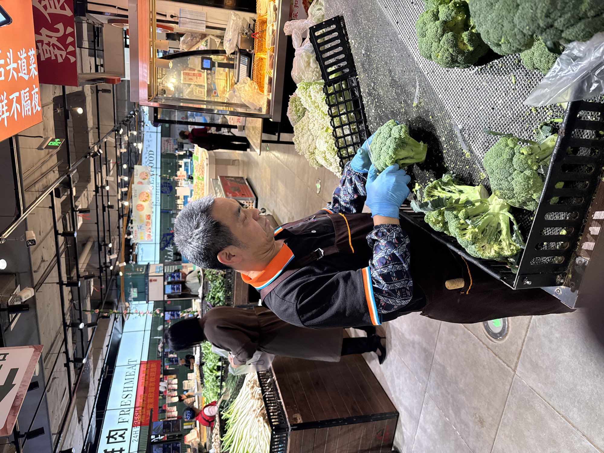An image taken in Jiangjin, China, shows a man in a supermarket wearing blue gloves sorting broccoli. He's wearing a black shirt with an orange-trimmed collar and patterned sleeves. He is placing broccoli into a black wire basket, and there are more broccoli heads on a mesh surface nearby. In the background, customers shop in the supermarket aisles, and signs for "FRESHMEAT" and "SEAFOOD" are visible. The floor is tiled in light gray. Another person is seen behind the man, with a light-colored purse in hand.