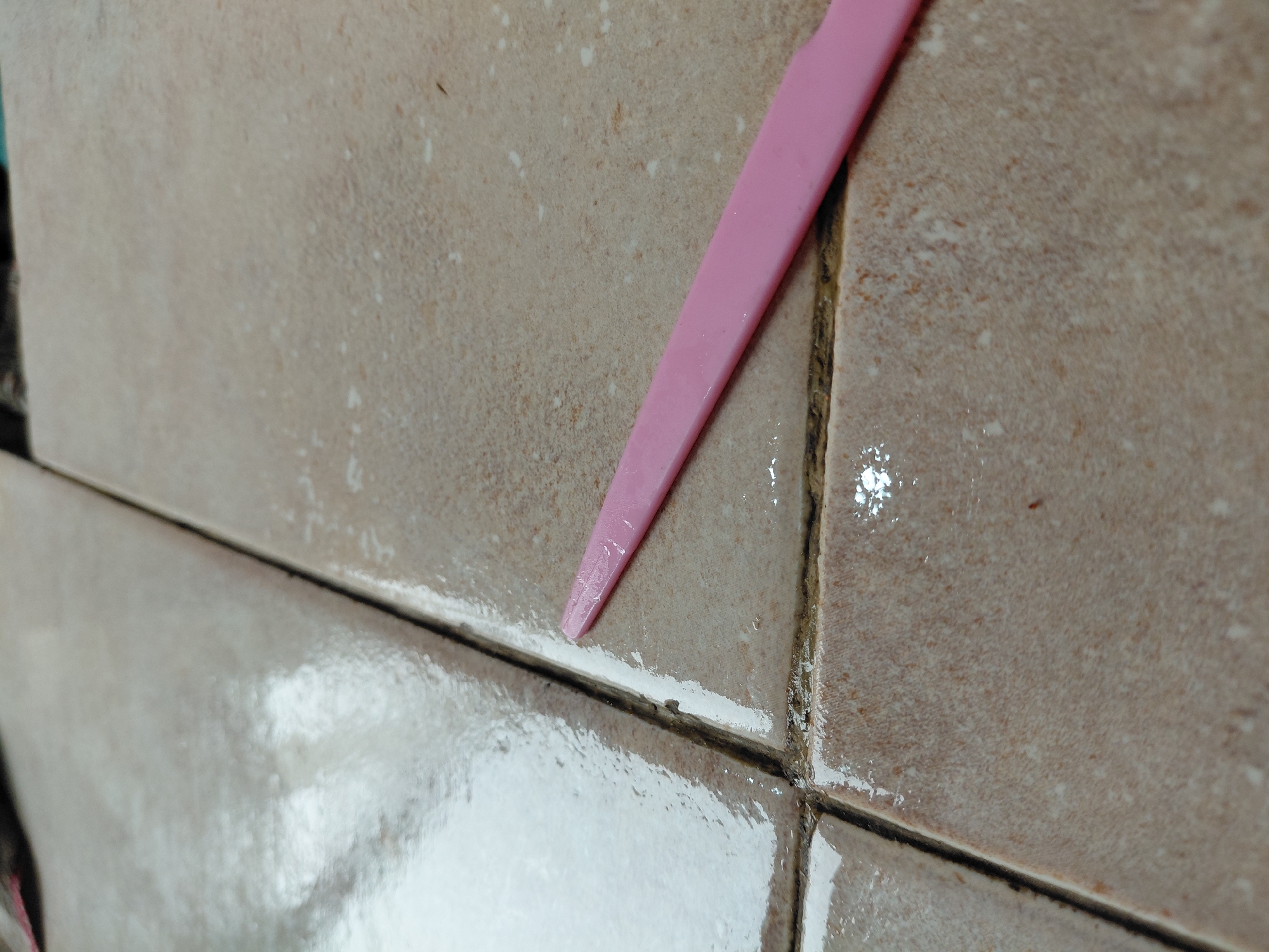The image displays a close-up view of a tiled surface, likely a wall or floor, within an indoor setting in Santa Ana, Venezuela. The tiles are a light beige or brownish color with a slightly textured finish. A bright pink, plastic tool, resembling a spatula or scraper, is inserted into a horizontal grout line, suggesting an ongoing process of cleaning, applying, or removing material from the grout.
The tiles and grout lines are wet and glistening, indicating the recent use of liquid, possibly water or a cleaning solution. The grout lines, particularly the darker vertical one on the right, show signs of discoloration or dirt. A white substance is visible along the horizontal grout line where the pink tool is situated, and also thinly spread on the edges of some adjacent tiles. This substance could be fresh grout, caulk, sealant, or residue from a cleaning product. The tool's action appears to be smoothing or scraping this material. No people or visible text are present in the image.