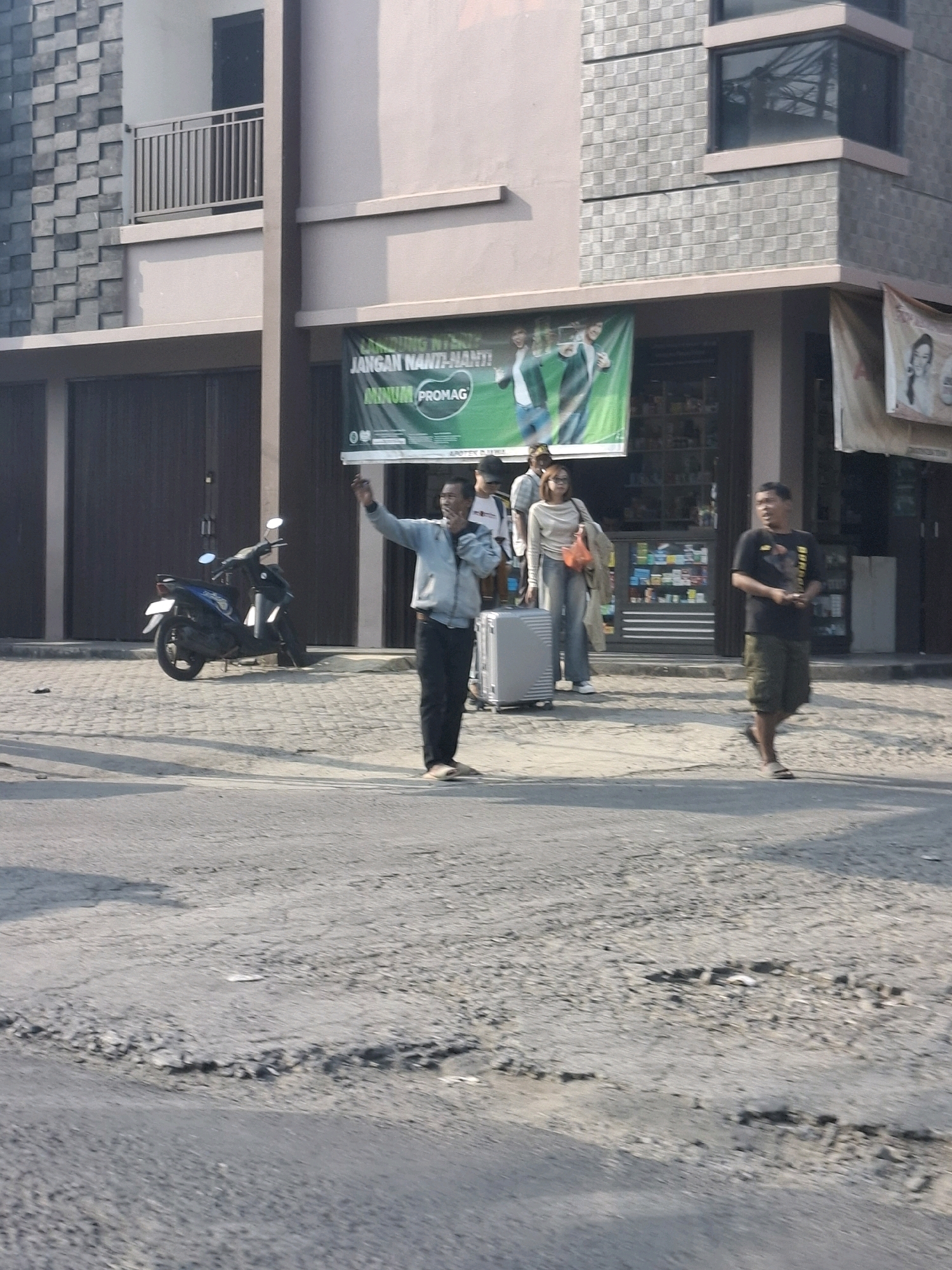 In Ciheulang Tonggoh, Indonesia, a group of people stand on a paved street in front of a multi-story building. A man in a light blue jacket and dark pants is in the foreground, gesturing with his right hand while holding a cigarette in his left. Behind him, a woman in a light-colored sweater and jeans walks with a rolling suitcase. To the right, another man in a black t-shirt and green shorts walks towards the foreground. A blue scooter is parked on the side of the street. The building behind the group has a large green banner with Indonesian text and images. The street appears to be in disrepair with visible cracks and potholes. The overall scene depicts a street-level view of an urban environment.