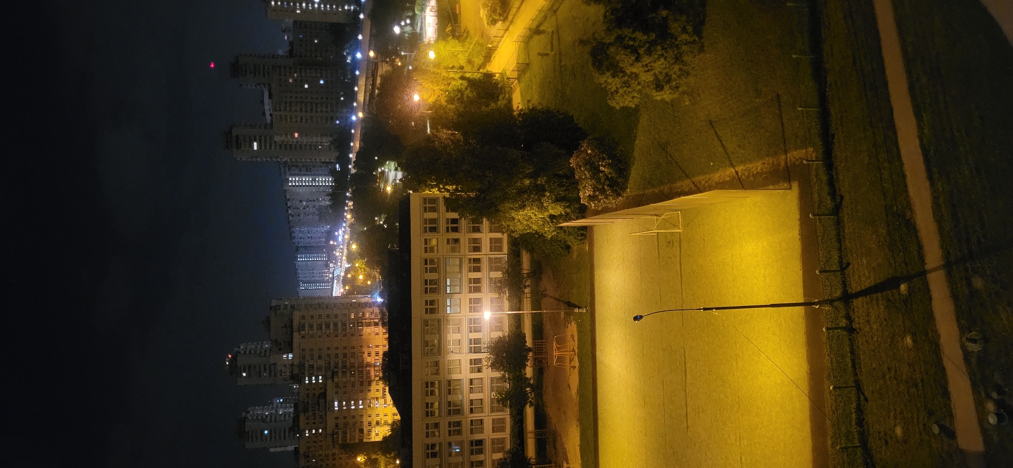 Here is a forensic description of the image:
The image presents an elevated, nighttime view of an urban environment. The left side of the frame features a dark sky contrasting with illuminated high-rise buildings, indicative of a city skyline. Streetlights punctuate the cityscape. To the right of the buildings are green areas and a park, bathed in artificial light, likely from floodlights. A building with multiple windows is visible in the foreground, also illuminated. A soccer field is present, marked by goalposts and intensely lit, contrasting with the shadowed periphery. A lamppost extends into the frame, casting a distinct shadow.