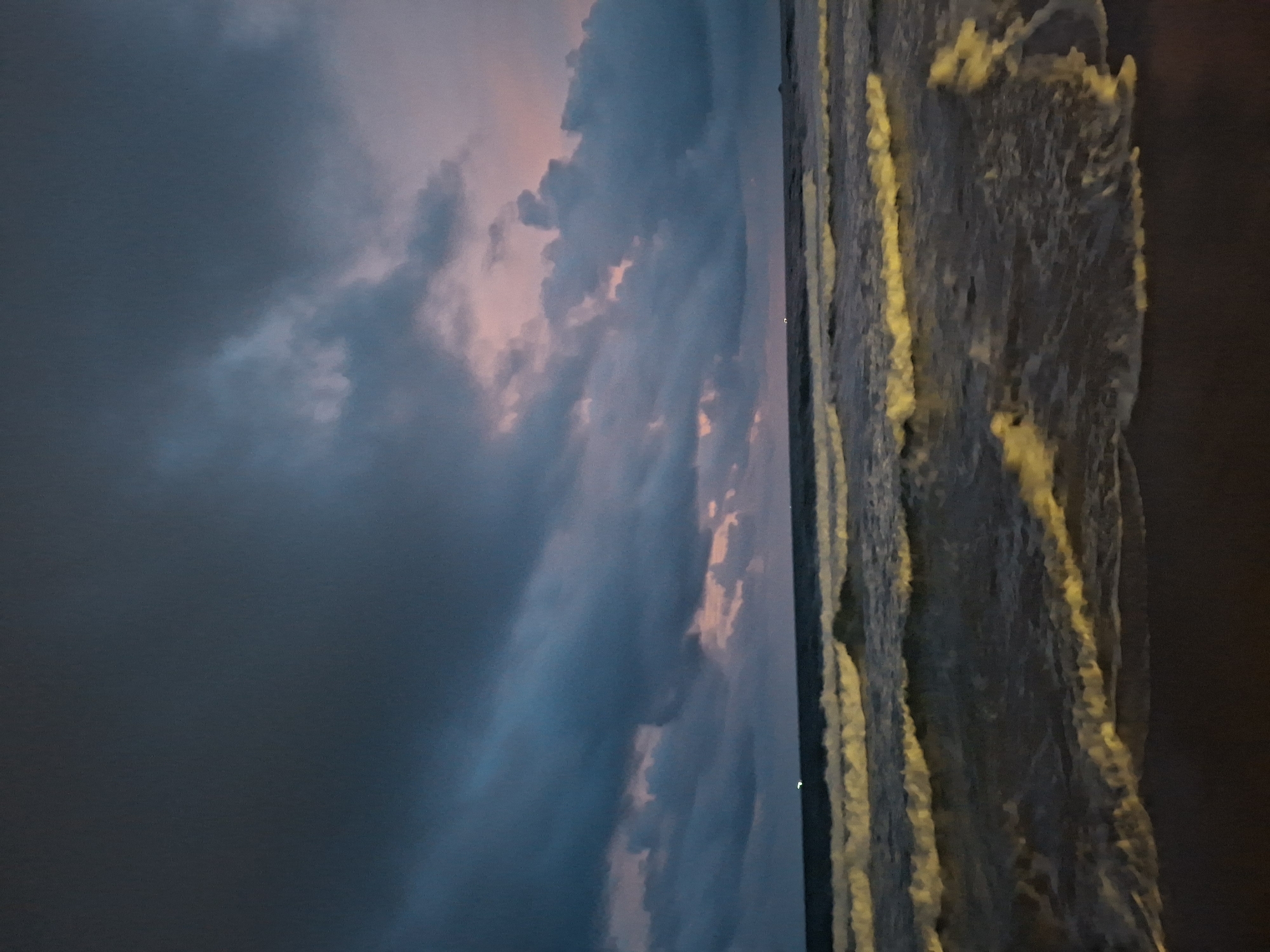 The image depicts a twilight scene at a beach, likely in Phú Quốc, Vietnam, characterized by dark, dramatic clouds streaked with pink and blue hues. The ocean is visible with multiple rows of waves breaking on the shore, their crests illuminated by an unseen light source, possibly artificial lights from the town or distant boats. The sand in the foreground is dark and appears wet, indicating the tide is near. There are no people or specific objects visible to suggest human activity beyond the implied presence of distant lights on the horizon. The overall atmosphere is somber and serene, capturing the transitional light of dusk.
