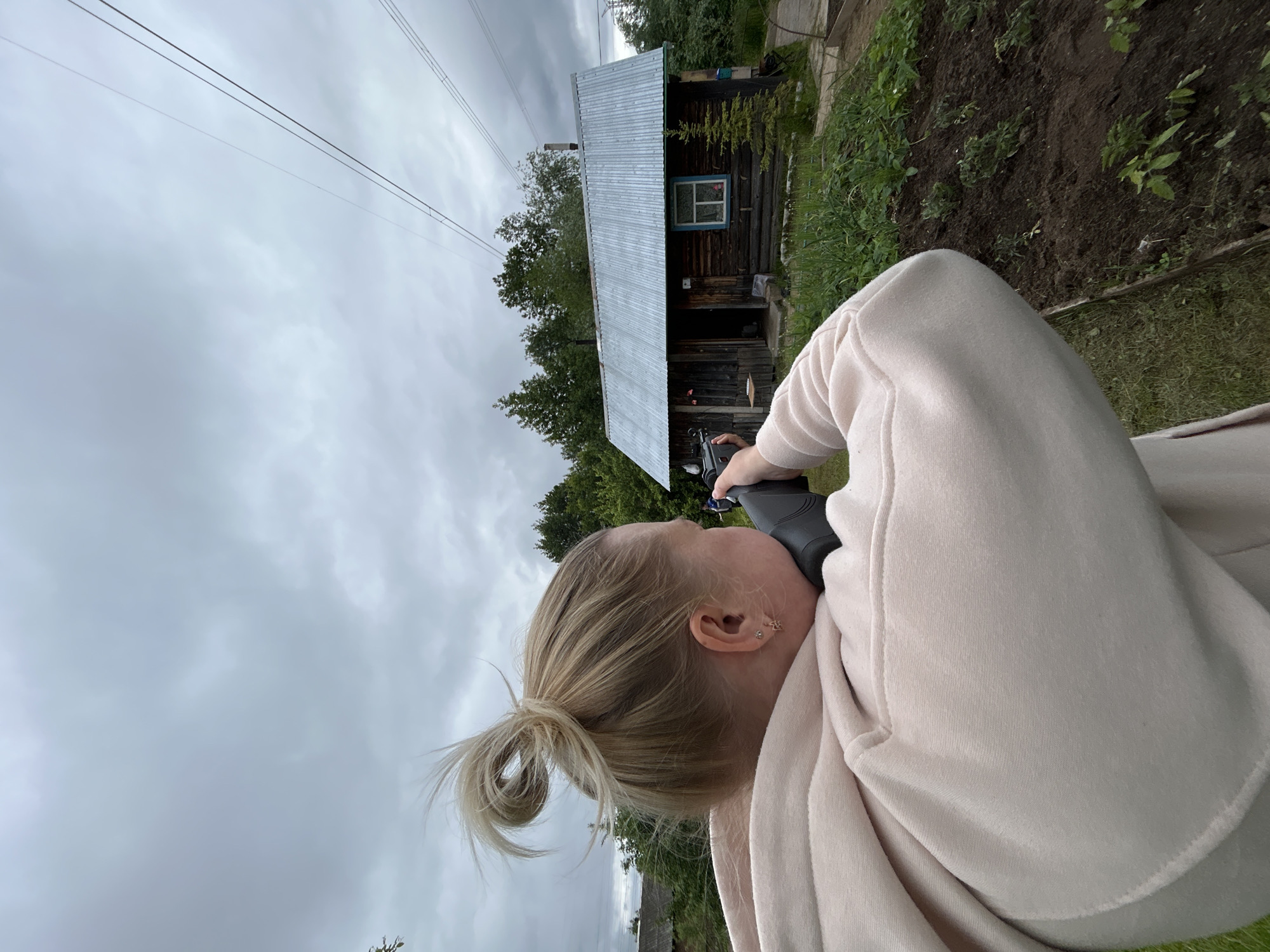 An individual with blonde hair tied in a messy bun, wearing a light-colored hooded sweatshirt, is depicted from a rear-right perspective in an outdoor setting in Chaikovsky, Russia. The person is holding a dark-colored firearm-like object, resembling a rifle or carbine, with its stock pressed against their right shoulder, in an aiming posture. The right ear is visible, adorned with two small earrings.
The target of the aim is a rustic wooden cabin in the mid-ground, constructed from dark logs and featuring a light blue window frame and a corrugated metal roof. A small coniferous tree stands adjacent to the cabin. Behind the cabin, dense green trees and foliage are visible. The foreground consists of green grass and a tilled garden plot with small plants. The sky is overcast with a uniform grey cloud cover. Two parallel power lines extend diagonally across the upper right portion of the frame.