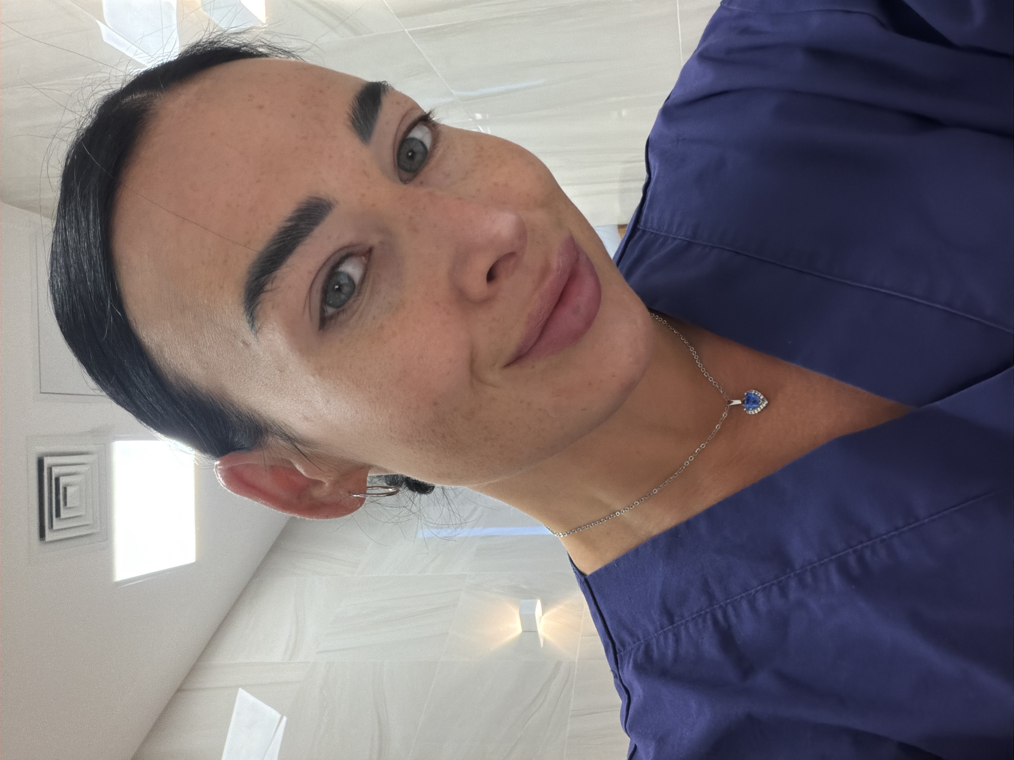 A woman is taking a selfie, angled slightly down. She has fair skin, dark hair pulled back from her face, freckles, and light-colored eyes. She is wearing a dark blue top and a silver necklace with a heart-shaped pendant that appears to have a blue stone. The background appears to be a modern bathroom with light-colored tiles and recessed lighting. The location is most likely not in Saint Petersburg, Russia, as the architecture and interior design elements are not typical of that city.