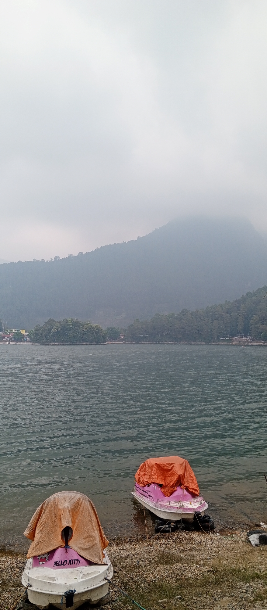 The image depicts a serene lake scene in Sarangan, Indonesia. Two small boats, covered with orange tarpaulins, are pulled up on a pebbled shore. One boat has a "Hello Kitty" decal. The lake extends towards a tree-lined shore, with a hazy mountain range in the background, suggesting an overcast or foggy atmosphere. The overall scene is calm and quiet, with no visible people.