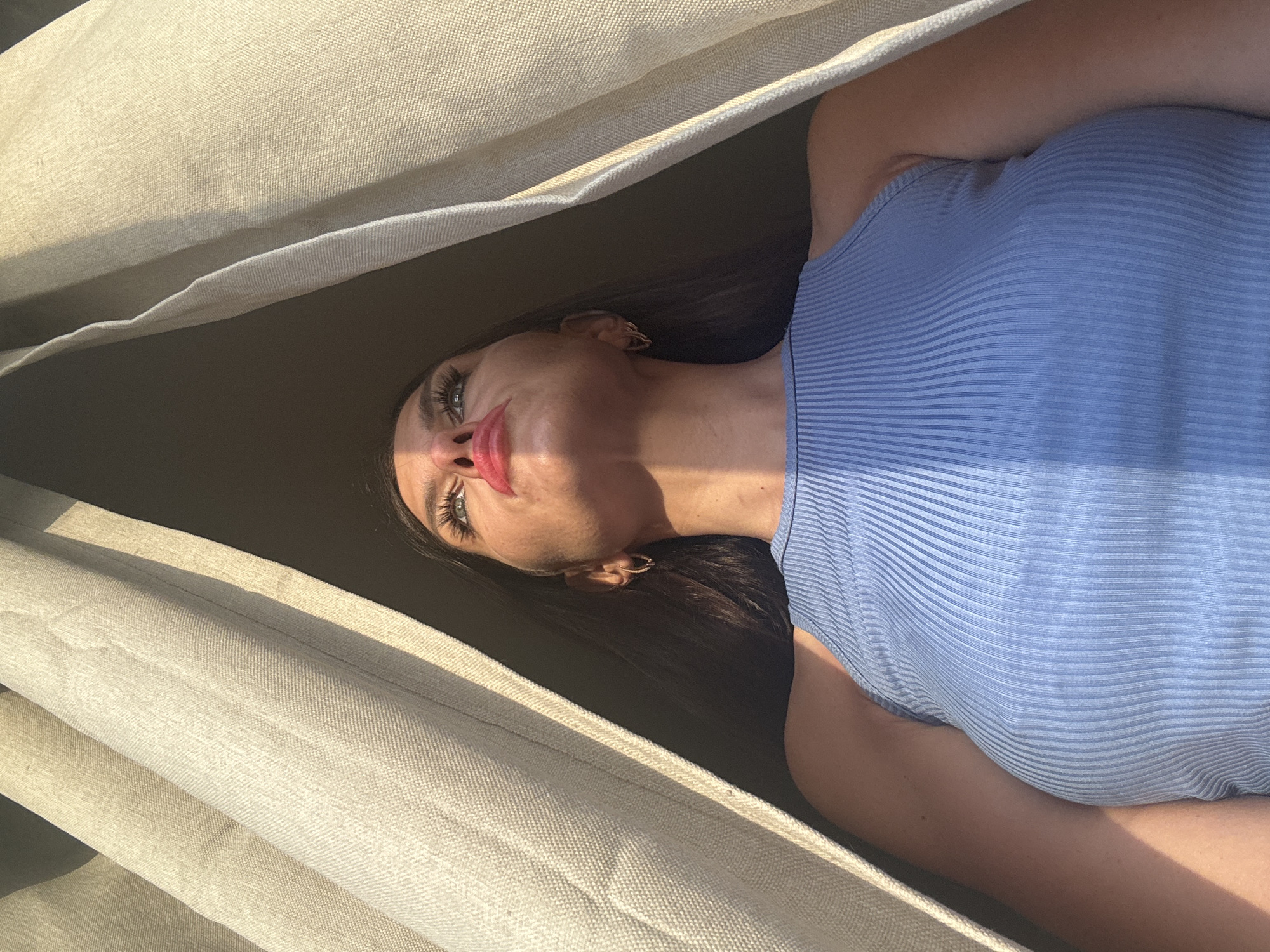 This is a vertical, eye-level, close-up, medium shot of a young woman lying down, possibly in a hammock, as indicated by the fabric surrounding her. She is looking upwards towards the camera. The woman has long, dark hair, striking green eyes, and full lips painted with a glossy, bright pink lipstick. She is wearing a light blue, ribbed, sleeveless top.
The scene is bathed in warm, natural sunlight, suggesting it is daytime, possibly late afternoon due to the long shadows and the golden hue of the light. The fabric around her appears to be a neutral, textured material, perhaps curtains or a canopy, creating a sense of intimacy and enclosure. The lighting casts a dappled effect on the fabric and her skin. The overall mood is relaxed and sensual. There is no visible text in the image.
The location context is Chaikovsky, Russia.