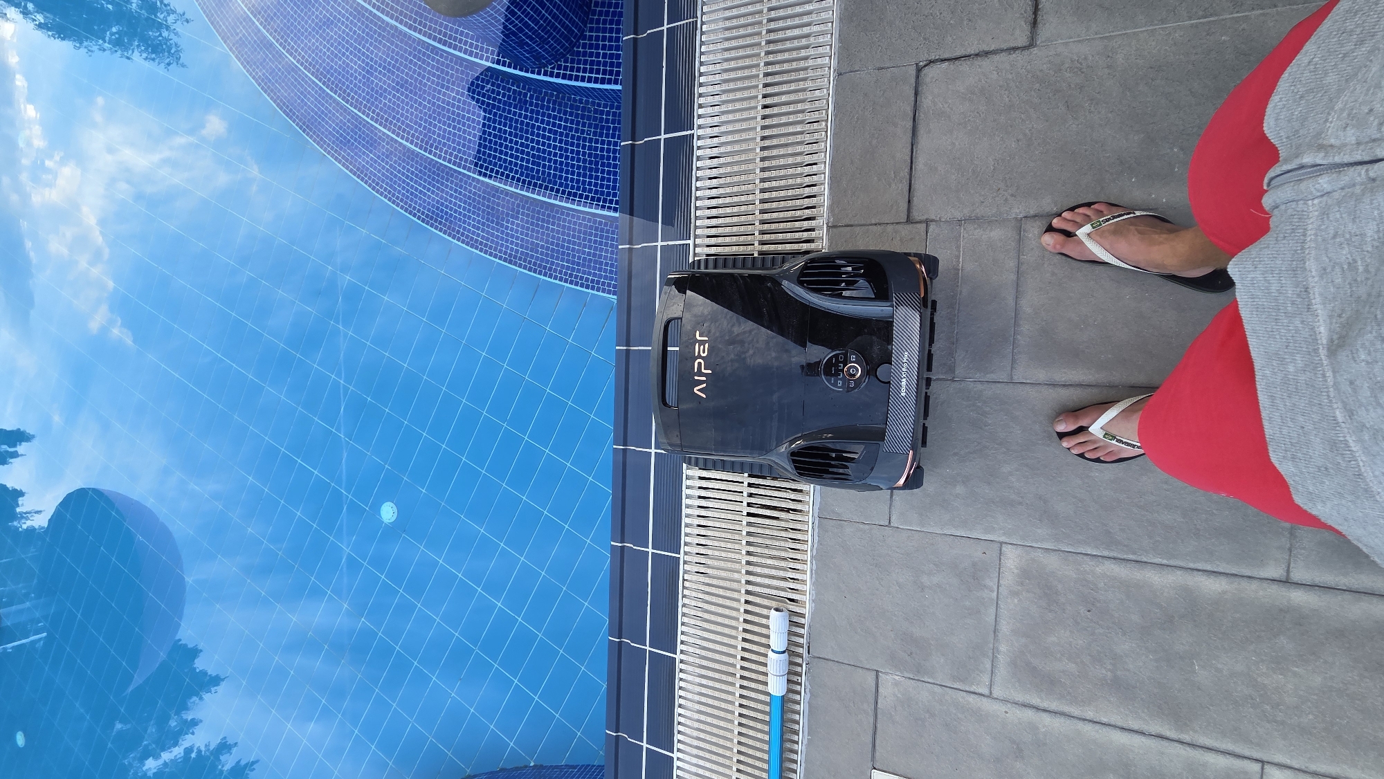 In an outdoor setting, a black robotic pool cleaner with "AIPER" logo is placed on a paved area next to a swimming pool. The pool is filled with blue water and reflects the sky and trees. There are tiled steps leading into the pool. A person's legs are visible in the foreground, wearing red shorts and flip-flops. The scene appears to be a sunny day.
