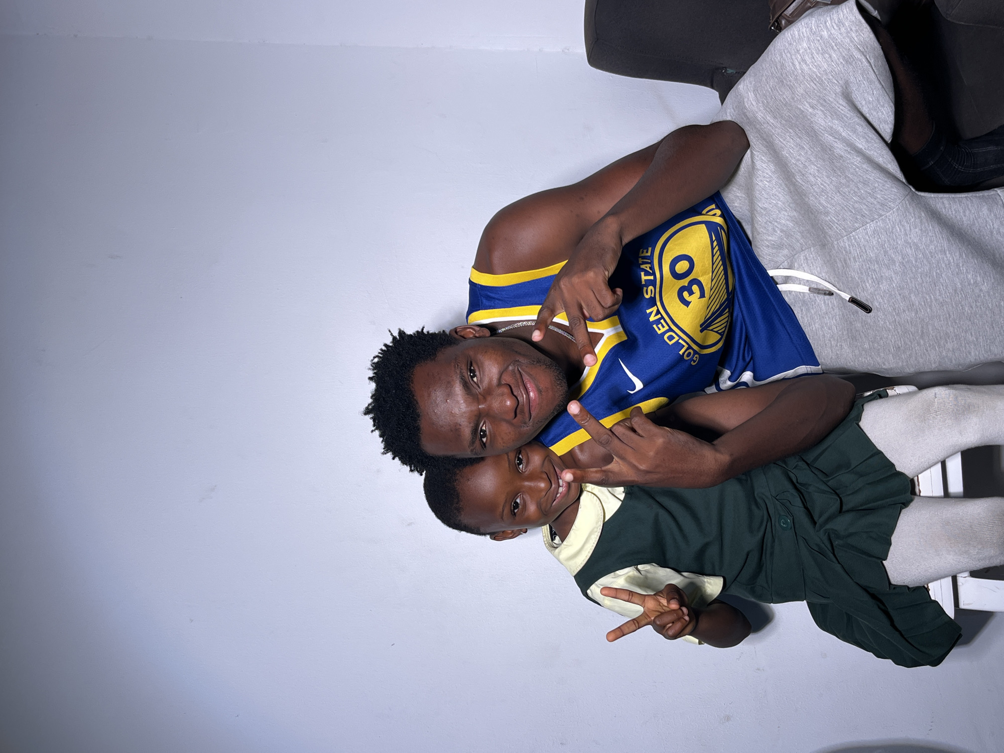 The image shows two people, an adult and a child, posed against a plain, light-colored background. The adult, on the left, is wearing a blue and yellow basketball jersey with the number 30 and the word "GOLDEN STATE" visible. They have their arm around the child. The child, on the right, is wearing a school uniform consisting of a light-colored collared shirt and a dark green pleated skirt and socks. Both individuals are smiling and making peace signs with their fingers. The adult has their other hand resting on their chest. The image is shot from a low angle, and the lighting is even, suggesting an indoor setting. There is no indication of the time of day or weather. The overall mood appears to be happy and affectionate.