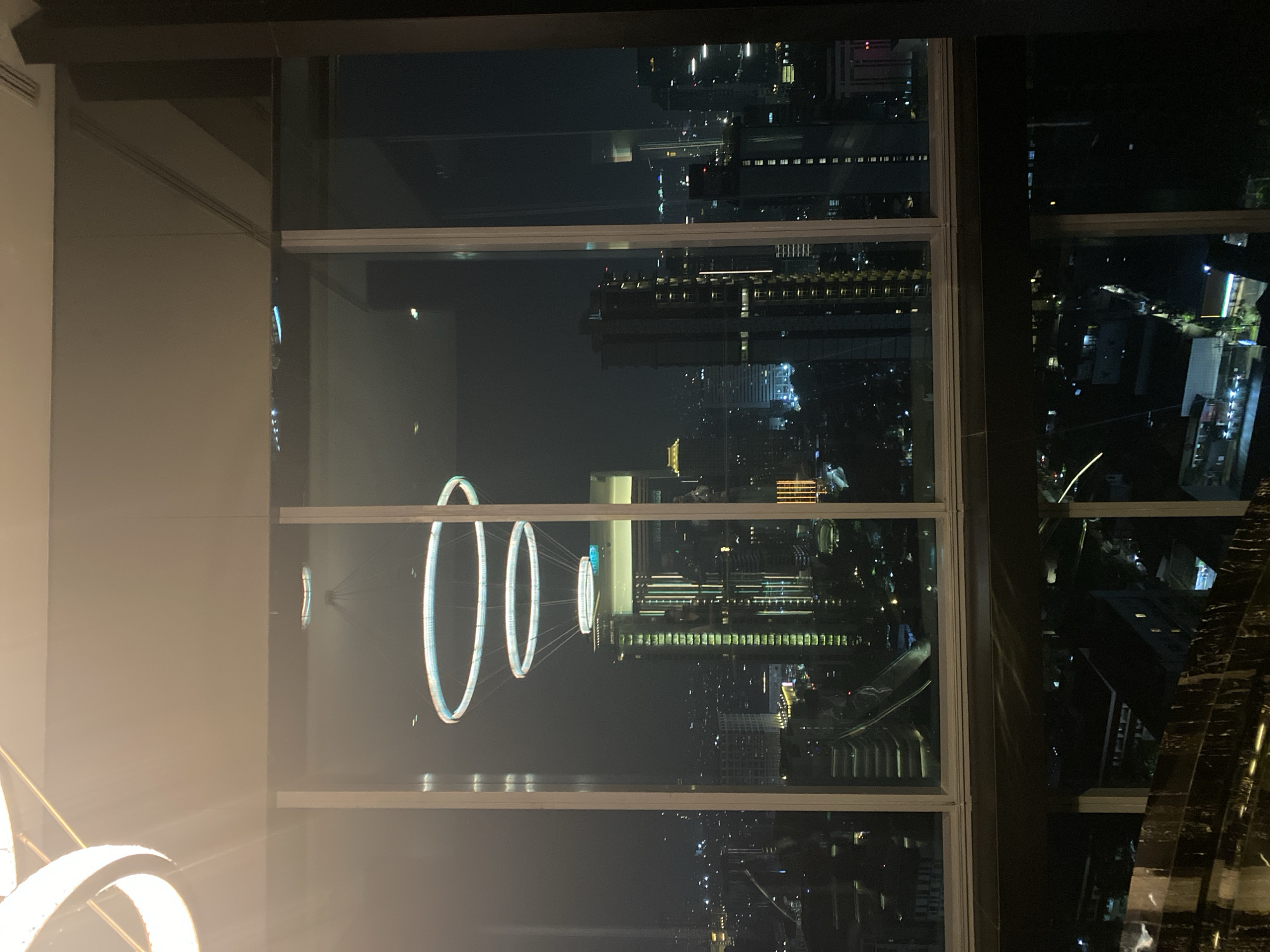The image shows a view of the city of Central Jakarta, Indonesia at night, as seen through a large window. The window has a dark frame and is divided into several panes. In the foreground, there are reflections of circular pendant lights, with one large light fixture visible in the top left corner. Outside the window, the cityscape is illuminated by numerous lights from buildings and streets, indicating a densely populated urban area. Several tall buildings, including skyscrapers, are prominent. The sky is dark, consistent with nighttime. In the lower part of the image, there is a reflection of what appears to be a dark, polished surface, possibly a table or railing, with some lights reflecting on it. The overall scene depicts a modern urban environment viewed from a high vantage point at night.