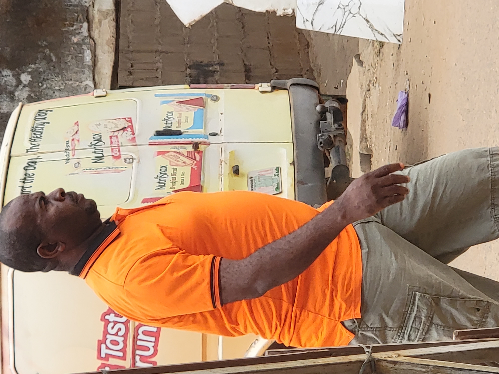 An adult Black man wearing an orange shirt and light-colored pants is positioned to the right of the rear of a light-colored van. The van displays advertisements for a product called "NutriSnax," including "Breakfast Biscuit" and the slogan "Start the Day, The Healthy Way" and "Tasty Run." The background shows an exterior environment, including building walls and a tan-colored ground surface. A purple wrapper lies near the rear of the van. The image's geographic origin is Maiburiji, Nigeria.