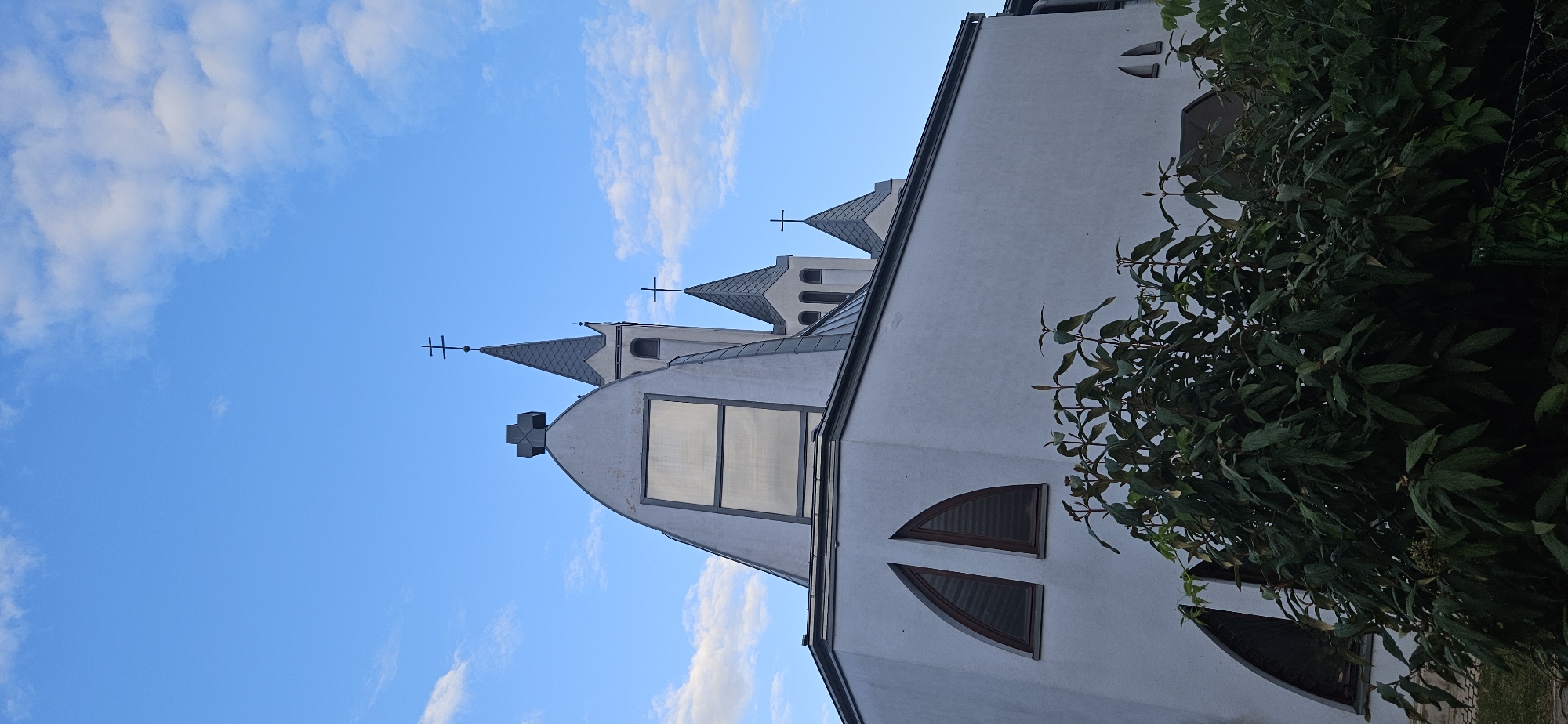 The image shows a modern white church in Hévíz, Hungary, against a blue sky with scattered clouds. The church has a distinctive architectural style with a curved roof and multiple spires topped with crosses. Two arched windows are visible on the left side of the building, partially obscured by lush green foliage in the foreground. The overall scene suggests a peaceful, perhaps early morning or late afternoon atmosphere, given the lighting. There are no people or animals visible in the frame.
