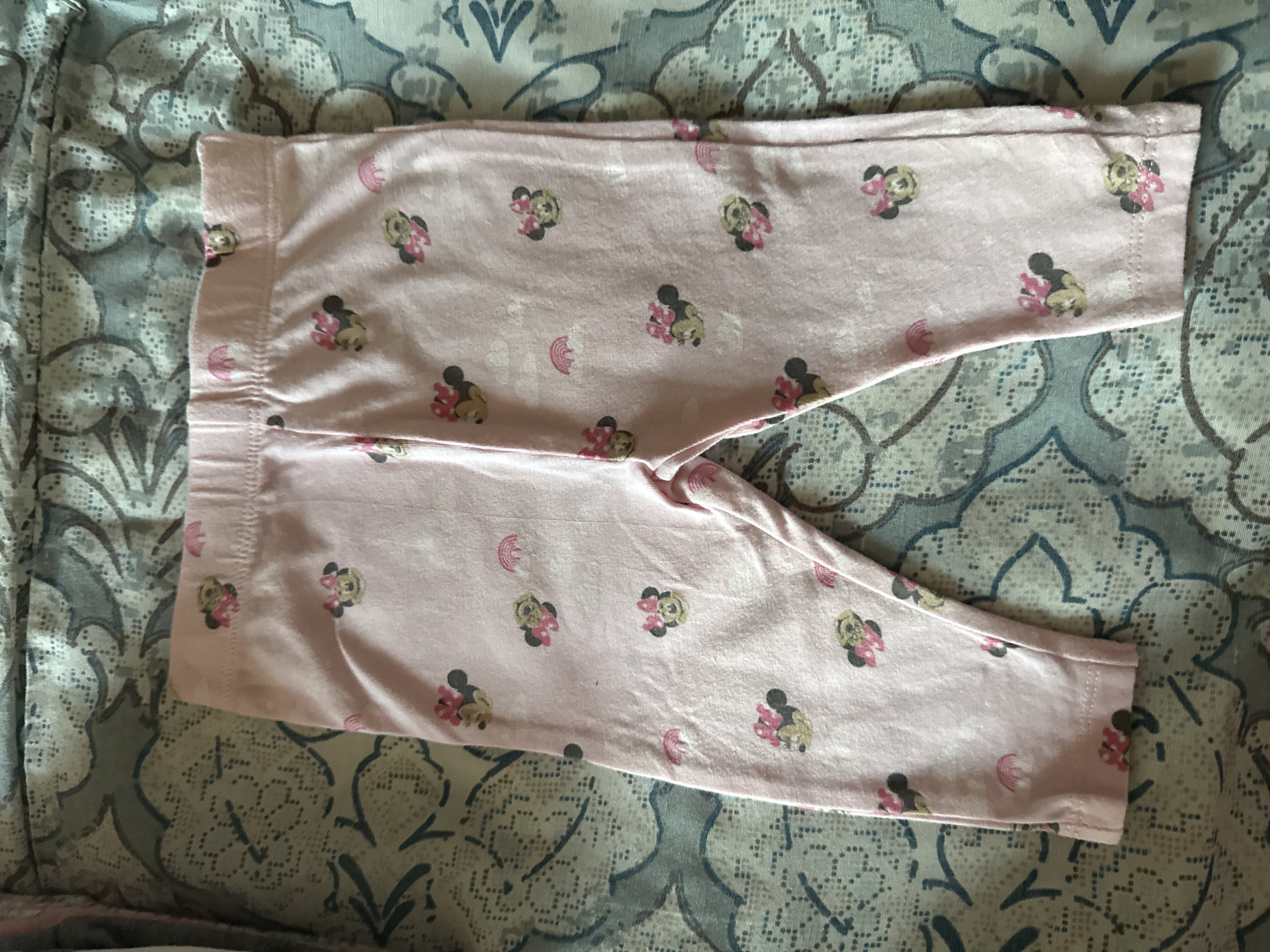 A pair of pink children's leggings with a repeating pattern of Minnie Mouse and small rainbows is laid flat on a patterned surface, which appears to be a bedspread or blanket with a blue and white damask-like design. The leggings are light pink and have a stretchy waistband. The Minnie Mouse graphics are small and show the character in various poses, some with pink bows. Small rainbow motifs are interspersed among the Minnie Mouse figures, along with what appear to be stylized clouds. The leggings are folded or creased in places, indicating they are not perfectly ironed. The overall impression is that of a piece of children's clothing casually displayed. No specific location in Tbilisi, Georgia is identifiable from the image.