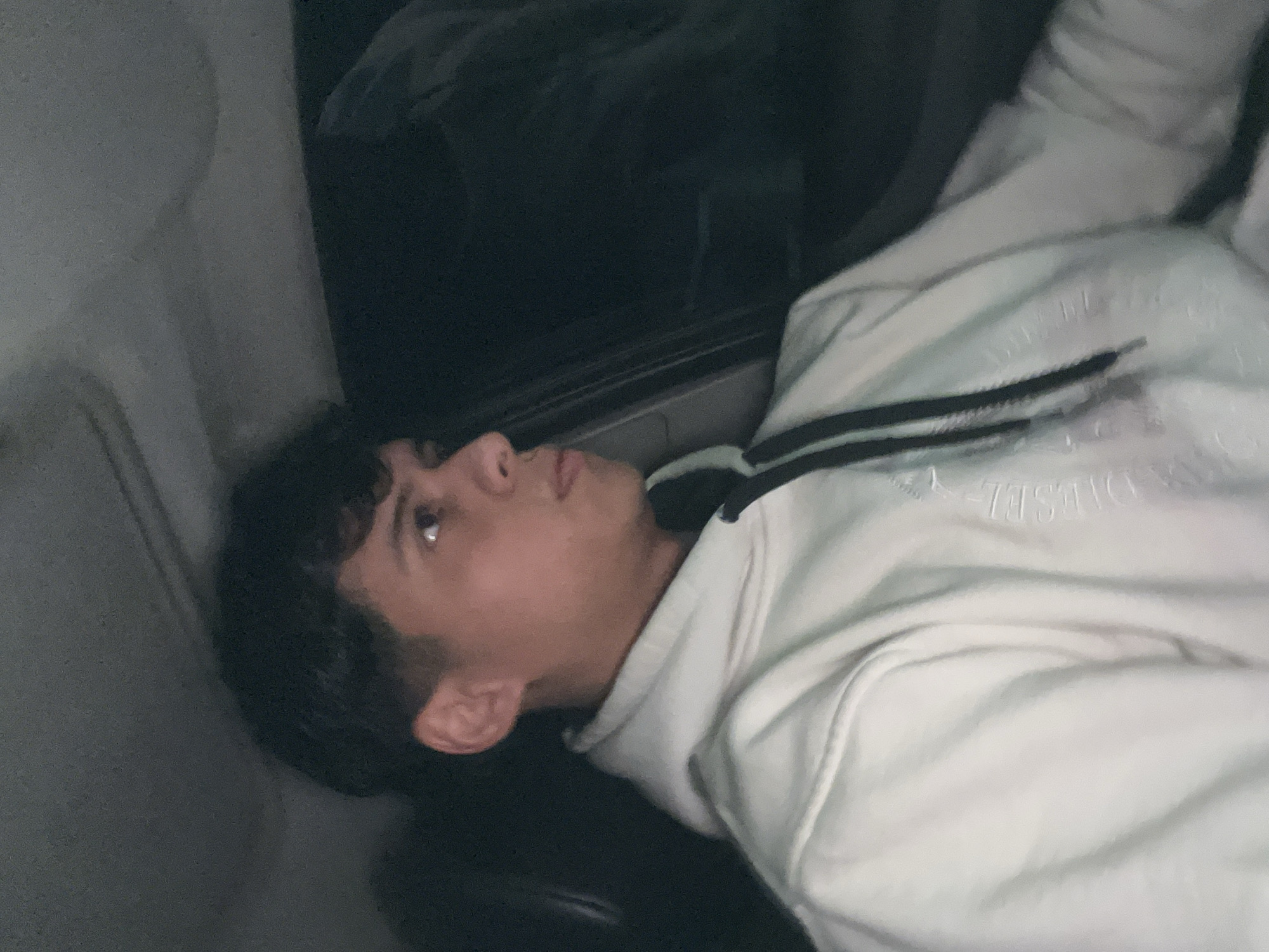 In an indoor, low-light environment, possibly a vehicle interior, a young male is depicted. He is seen from the chest up, turned to his right, with his face angled away from the camera and his gaze directed off-frame. He has dark, wavy hair and is wearing a light-colored hooded sweatshirt with black drawstrings. The sweatshirt has a subtle embossed pattern or logo. The background is dark and out of focus, suggesting a night-time or dimly lit setting. The image does not provide any specific visual cues to determine the location in San Fernando del Valle de Catamarca, Argentina.