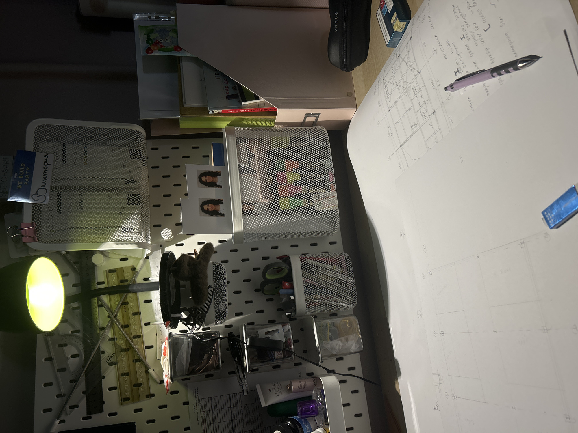 This image shows an organized workspace. A bright lamp illuminates a desk covered with a large sheet of paper containing architectural drawings and handwritten notes in Bulgarian. A lavender-colored pen rests on the paper. To the side is a collection of storage containers mounted on a pegboard, holding items like rulers, a stuffed animal, scissors, pens, and toiletries. A file organizer sits on the desk, along with a black Vogue-branded pencil case and a pack of cigarettes. There are also two photos of a female person visible on the pegboard. The OCR of the image includes street names, measurements and architectural notes in Bulgarian, suggesting that the architectural drawing on the desk is for a building in Sofia, Bulgaria.
