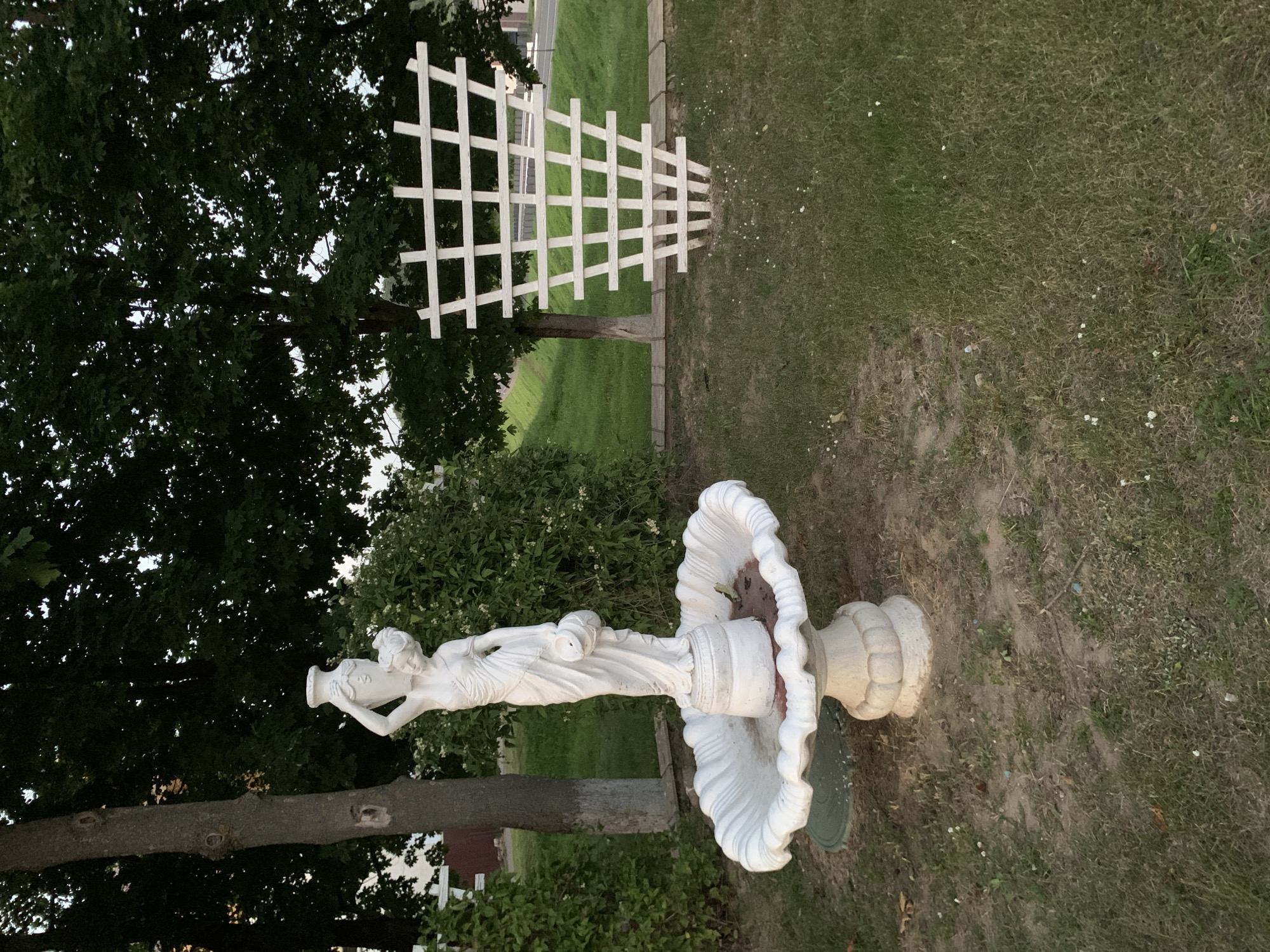 The image captures a view of a garden setting in Kapyĺ, Belarus. In the foreground, a white statue of a woman carrying a vase on her head stands within a shell-shaped fountain. The statue is made of a material resembling plaster or concrete. Behind the statue, a white wooden trellis, shaped like an inverted pyramid, is positioned on the lawn. The area is surrounded by lush green grass, mature trees with dense foliage, and some bushes. A paved surface and a road are visible in the background, suggesting an urban or suburban environment. The overall scene depicts a well-maintained outdoor space with decorative elements.