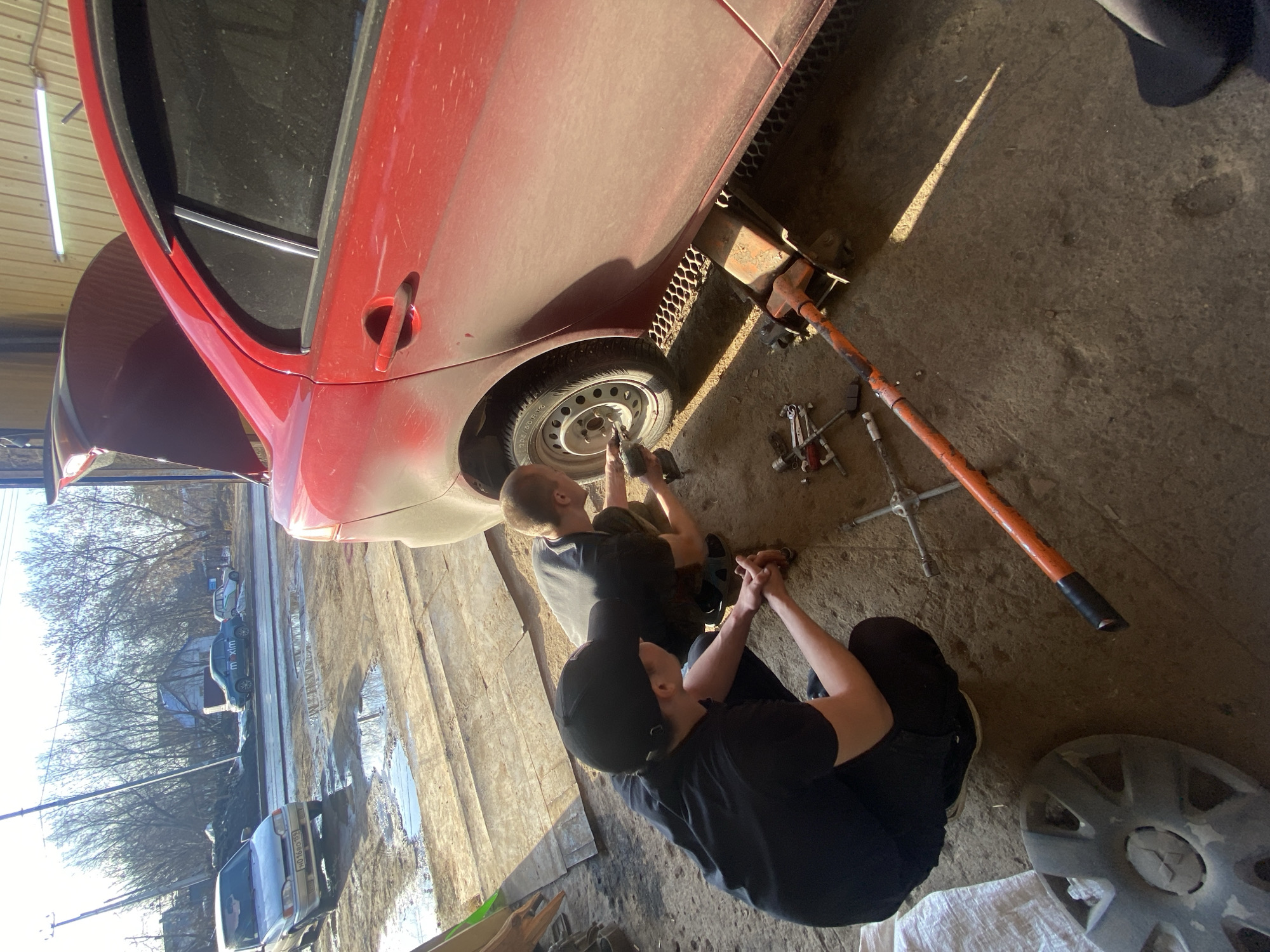 Two men are working on a red car in what appears to be a garage or workshop. The car is elevated by a jack, and one of the men is using a power tool on the wheel. The other man is sitting nearby, watching. Various tools and car parts are scattered around the floor. Outside, beyond the open garage door, the scene includes trees without leaves, puddles of water, and parked cars, suggesting a street view possibly in Chaikovsky, Russia. One of the vehicles has the name "maxim" on it.