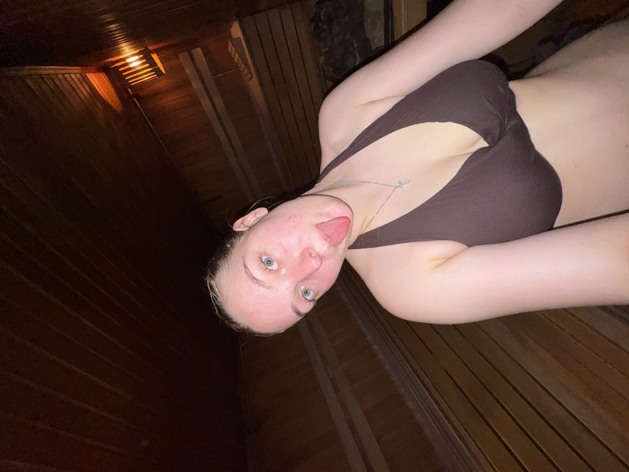 A woman with fair skin and blue eyes is posing for a selfie in what appears to be a sauna. She is wearing a brown bikini top and a silver cross necklace. Her tongue is sticking out. The interior walls of the sauna are wood, and there is a dim light fixture visible. No information about the location in the city of Chaikovsky, Russia, is discernible.